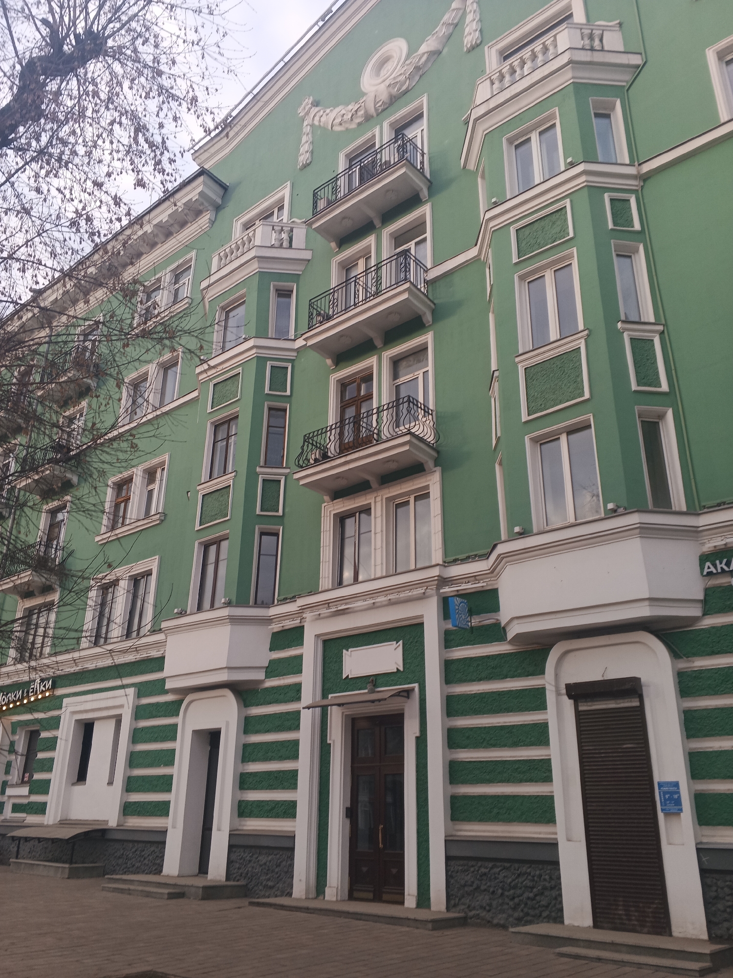 The image shows a green building in Perm, Russia, with white architectural details, including window frames, horizontal stripes, and decorative elements. The building has multiple stories with balconies on some floors. The facade features different types of windows, some framed with textured surfaces and others with plain surrounds. At street level, there are several entrances, including one with a closed shutter. A sign in Cyrillic is visible above one of the entrance areas. The image suggests an urban environment, with a sidewalk and trees visible in the foreground. The sky is overcast.