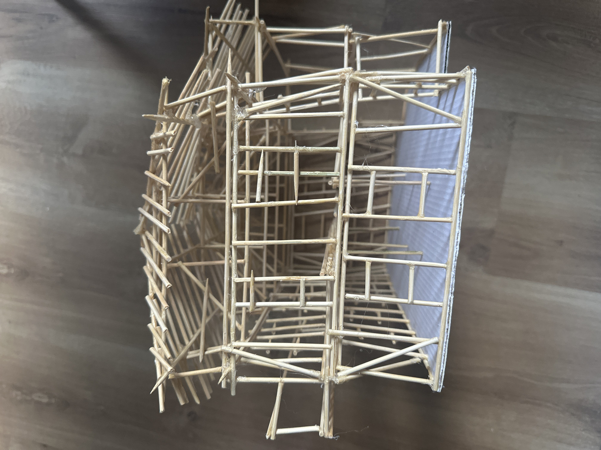The image shows a handmade model of a building, possibly architectural, constructed primarily from thin wooden sticks, resembling bamboo skewers or similar materials. The model exhibits a complex structure with various intersecting and overlapping elements. Some sections appear to be partially covered with a translucent white material. The model is resting on a brown, wood-grained surface, likely a tabletop or floor. There is no information about the location being in Sofia, Bulgaria, or any other specific geographic location. No people, subjects, actions, or interactions are visible in the frame. The environment is limited to the immediate surface on which the model rests.