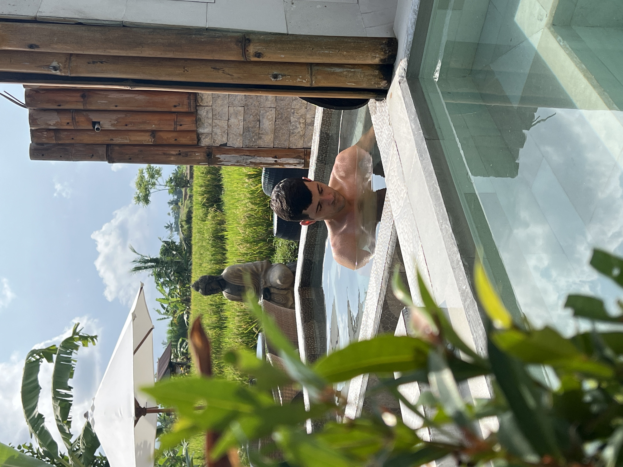 The image depicts a man partially submerged in a rectangular pool of water, likely in Singakerta, Indonesia, given the presence of rice paddies in the background. The pool has a concrete or stone border and reflects the sky's clouds. A bamboo structure is visible behind the pool, and a statue of a Buddha is situated on a grassy area. Green foliage is in the foreground.