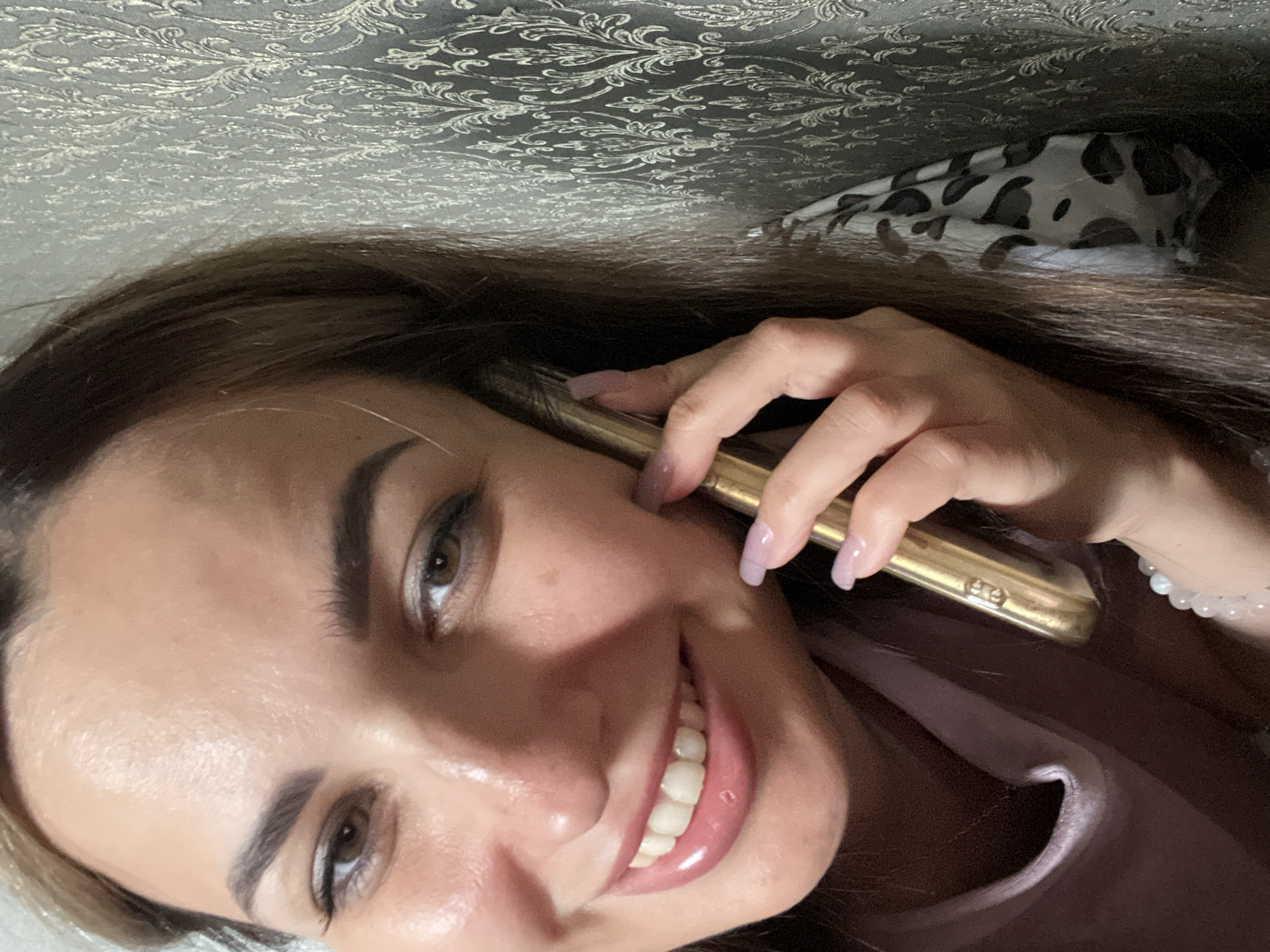A young woman with brown hair and fair skin is holding a gold smartphone to her right ear, talking on the phone. Her eyes are brown and she is smiling. She is wearing a light pink shirt. The background is a patterned wallpaper. The image is a close-up and the location is unknown.