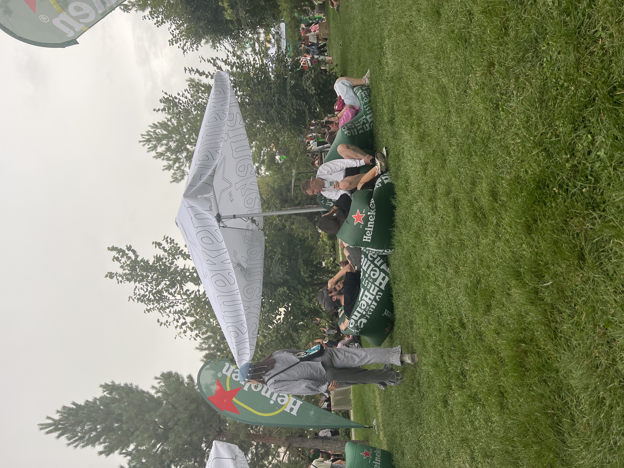 This image depicts an outdoor event at Pervomayskiye Prudy, Kazakhstan, with a focus on a Heineken-branded area. Several individuals are seen lounging on large, green, inflatable beanbag chairs. One person, a woman in a grey checkered outfit and a blue cap, walks away from the camera towards the left of the frame. Others are seated or reclining on the beanbags, some engaged in conversation and others looking at their phones. A large white umbrella with Heineken branding provides shade over some of the seating. In the background, trees form a natural backdrop, and more people and structures, possibly stalls or stages, are visible, suggesting a larger festival or gathering. The ground is covered in lush green grass. A tall, green flag with a red star and Heineken branding is prominently displayed on the left side of the image. The overall atmosphere appears relaxed and social.