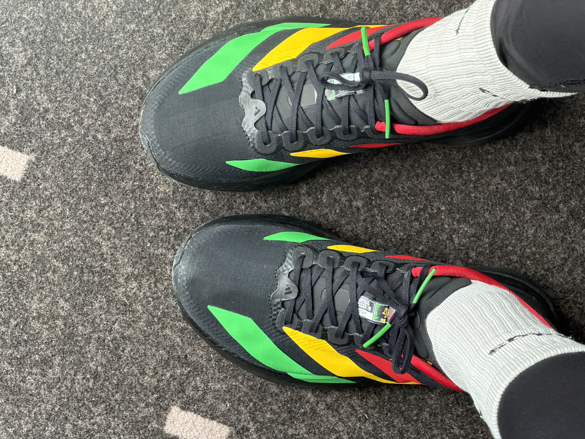 The image shows a pair of black Adidas running shoes with red, yellow, and green accents, worn with white socks and black pants. The shoes are positioned on a dark gray carpeted surface. The perspective is from above, looking down at the shoes and feet. The scene is indoors, as indicated by the carpet and lighting. The location in the city of Bremen, Germany is not discernible from the provided image.