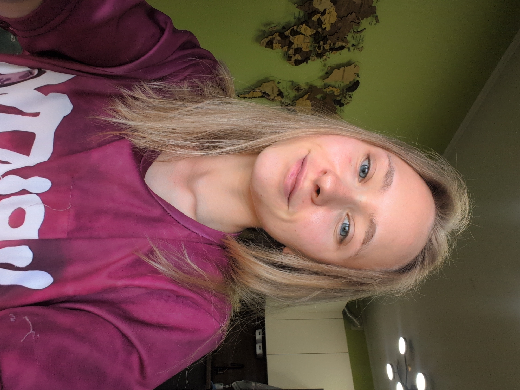 A young Caucasian woman with blonde hair and blue eyes is taking a selfie in a room with a green wall. She is wearing a purple t-shirt with white text on it. A wooden world map is mounted on the green wall behind her. The image appears to have been taken indoors.