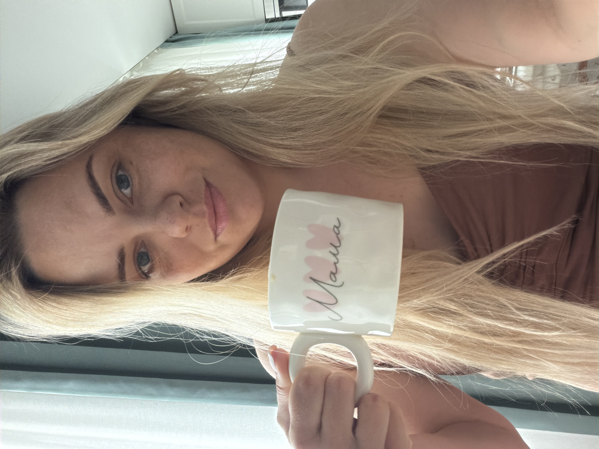 A blonde-haired woman is holding a white mug with the word "Mama" written on it in a handwritten-style font with pink floral accents. She is wearing a brown top. The environment appears to be indoors, with a window visible in the background, suggesting a room in a house or apartment. The location is not discernable to be Votkinsk, Russia.