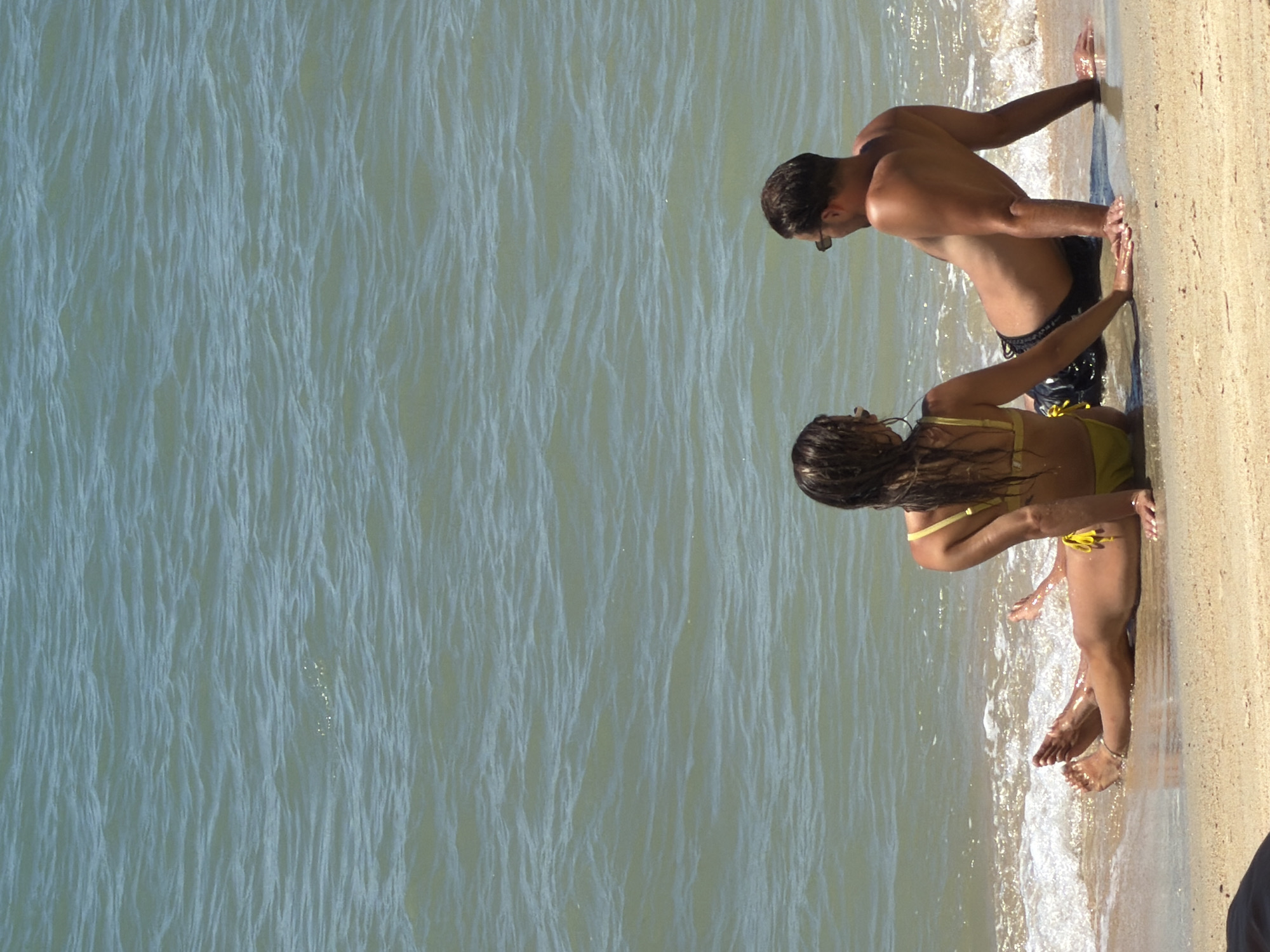 Two individuals are sitting on a sandy beach in Baan Mae Nam, Thailand, with their backs to the camera. The person on the left, appearing to be female, is wearing a yellow bikini and has long, dark hair. The person on the right, appearing to be male, is wearing dark swim trunks and sunglasses. Both are facing the ocean, which has gentle waves lapping at the shore. The water is a light blue-green color. The sand is a light beige color. The image captures a moment of relaxation by the sea.
