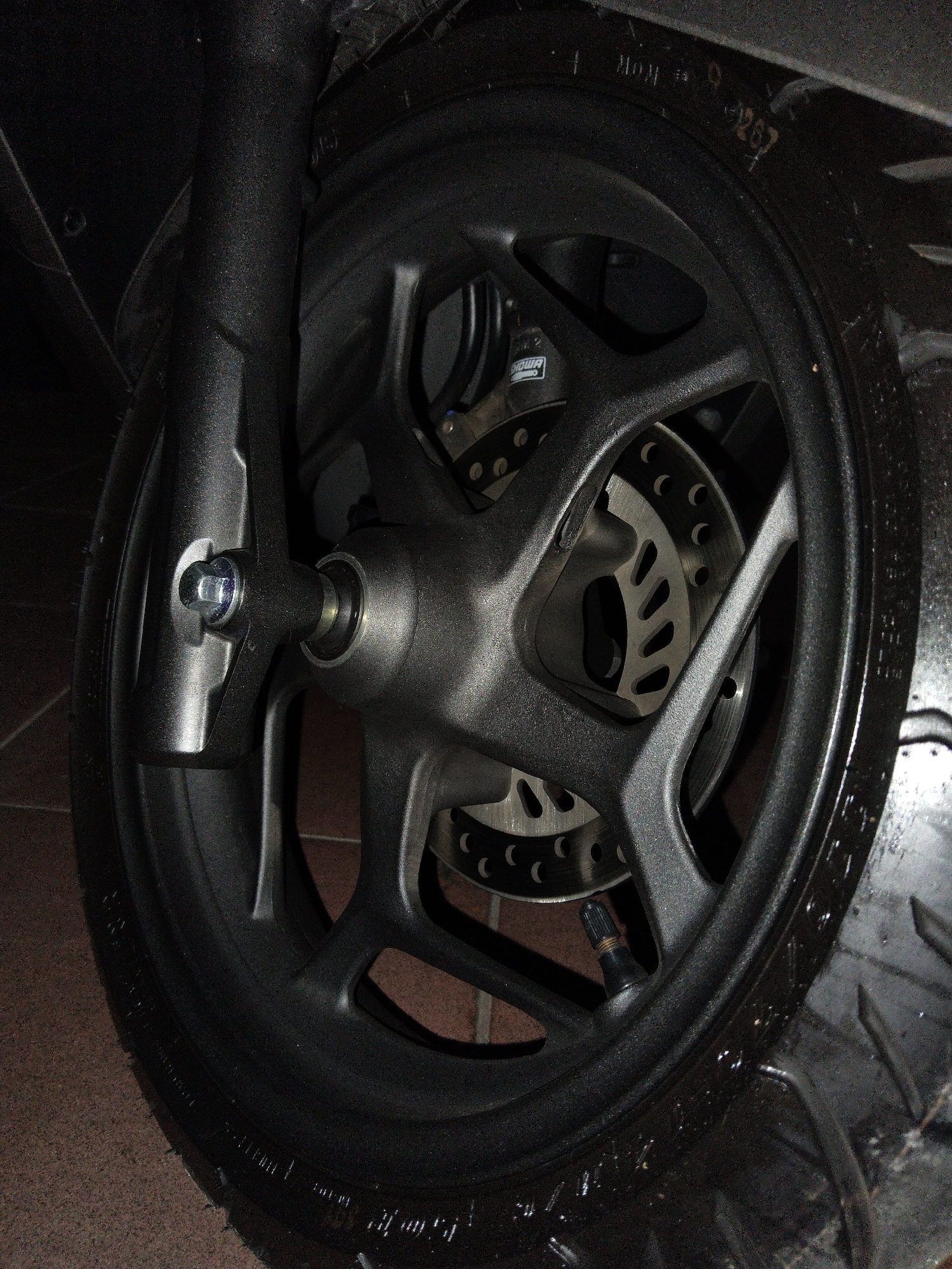 A close-up, low-angle, full-frame shot shows the front wheel and brake assembly of a dark-colored motorcycle. The motorcycle is parked on a reddish-brown tiled surface, suggesting an indoor location, possibly a garage or workshop, in Banyurojo, Indonesia. The wheel is black with a multi-spoke design. A perforated brake disc is visible behind the spokes. The front suspension fork is black and extends upwards from the wheel hub. A silver bolt is visible securing the wheel to the fork. The tire is black and shows some reflections, indicating it may be wet or polished. Some markings are visible on the tire sidewall. The scene is dimly lit, with highlights on the metallic parts of the wheel and fork.