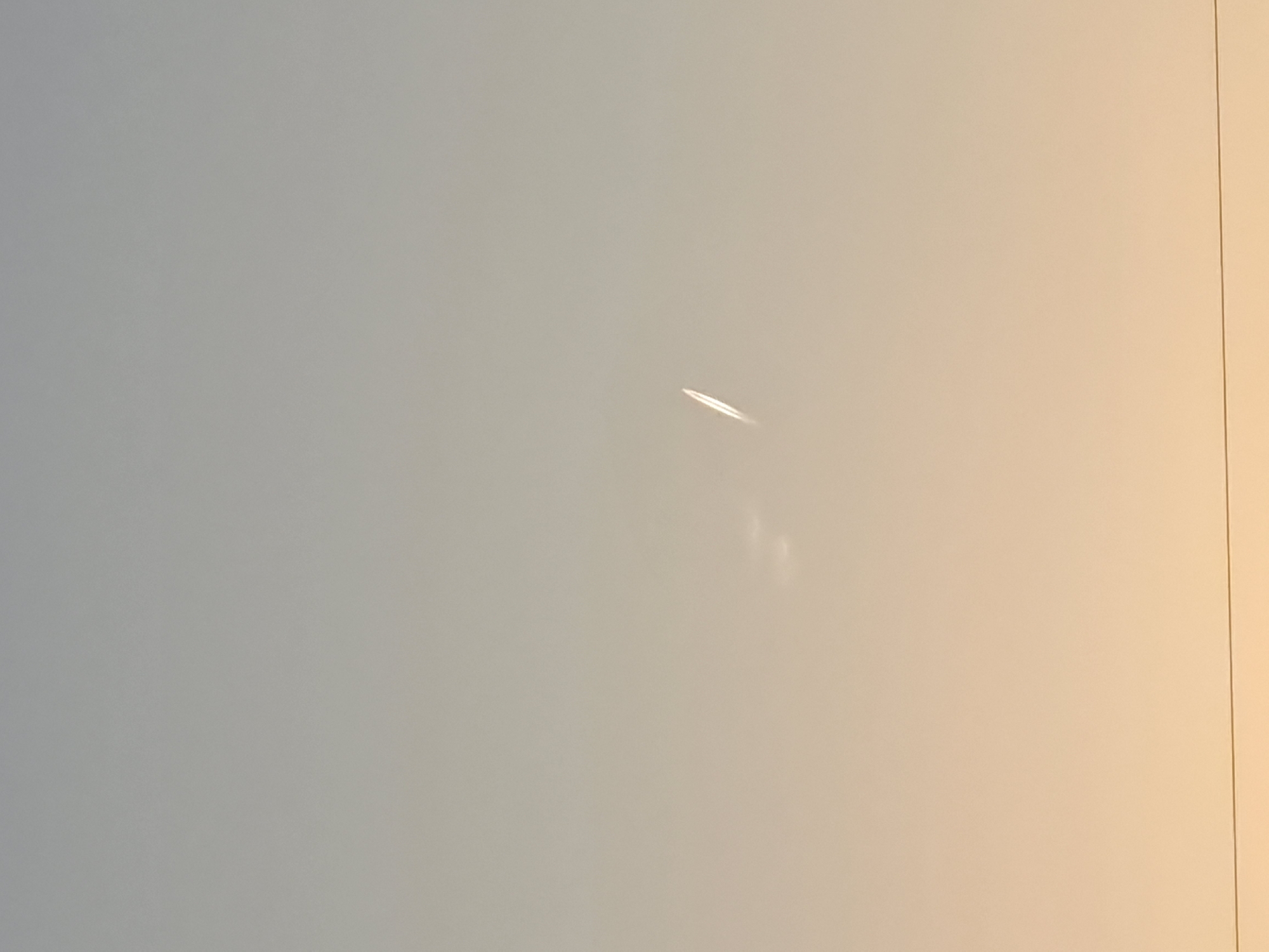 The image shows a contrail in a light-blue and slightly peach-colored sky. The contrail is short and bright. The right edge of the image suggests the edge of a white or light-colored wall or object. There are no people, subjects, or objects other than the contrail and the indeterminate white surface. The environment is essentially the sky. The location is unknown. There is nothing in the image to indicate that it is in Negovan, Bulgaria.