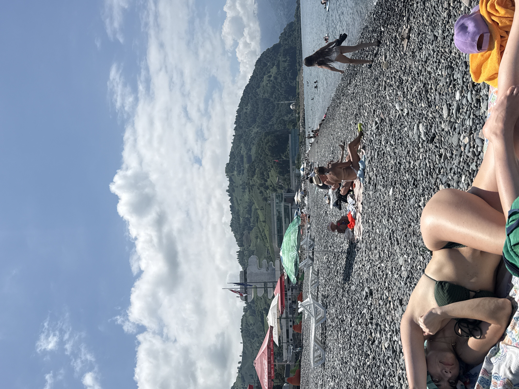 The image depicts a sunny day at Sarpi beach, Georgia. In the foreground, a woman in a green bikini lies on a towel, her head resting on her arm. She wears a blue headscarf. The beach is covered in dark grey pebbles. In the mid-ground, several beach umbrellas are set up, some red and one green. White lounge chairs are scattered on the beach. Further back, people are seated on towels and some are in the water. A young child sits on a towel near a green umbrella. In the background, lush green hills rise behind the beach, with a building featuring flags visible on one of the slopes. The Black Sea stretches out to the right of the image, with several people swimming or on paddleboards. The sky is blue with scattered white clouds.