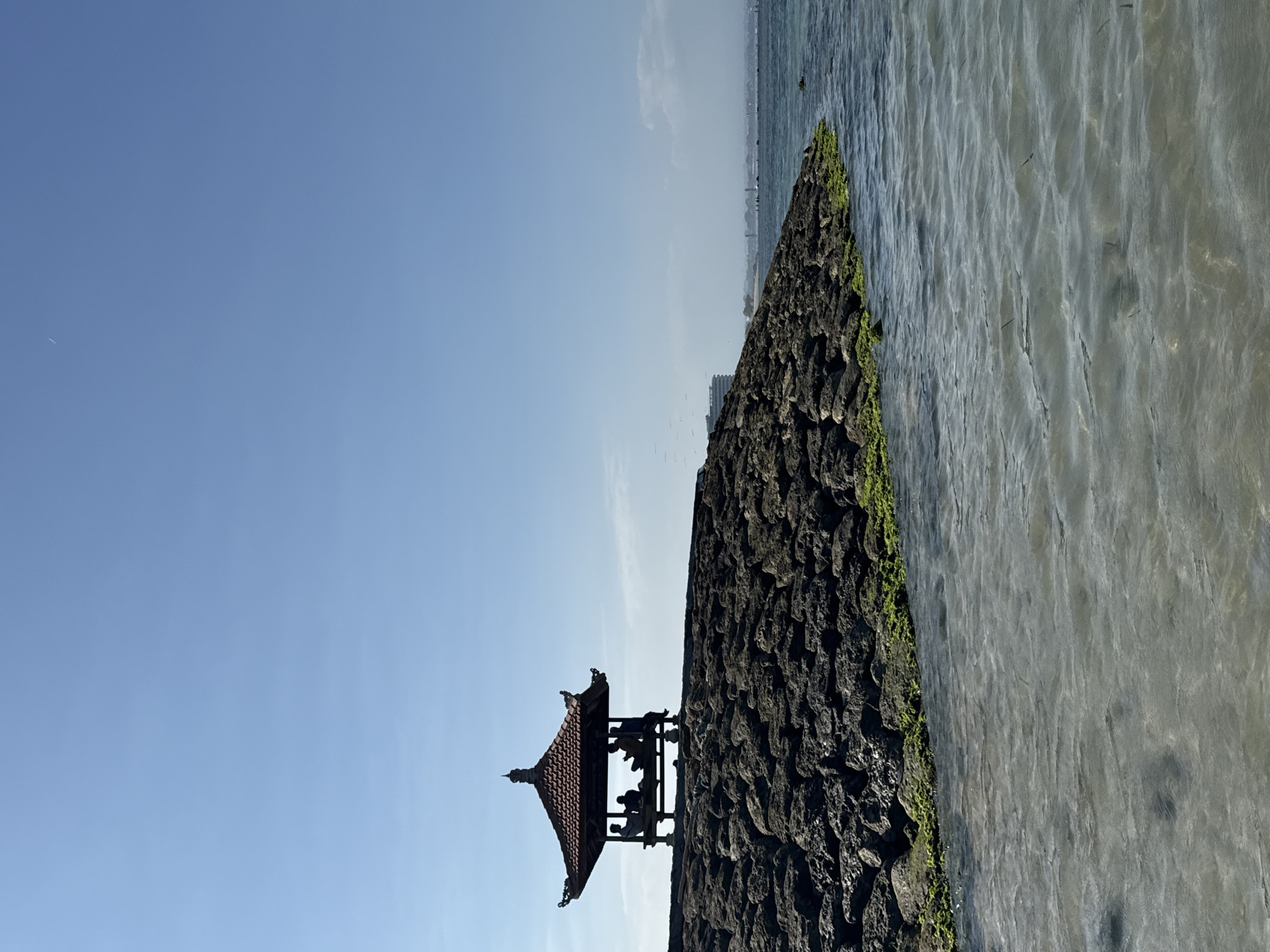 A serene tropical scene unfolds on a sunny day in Denpasar, Indonesia. In the foreground, the clear, shallow water of the ocean gently laps against a rocky shoreline. The rocks are partially covered in vibrant green moss, suggesting a humid, coastal environment.
Dominating the mid-ground is a breakwater constructed from large, dark, irregular stones. Perched atop this breakwater is a traditional Balinese gazebo, its tiered, terracotta-tiled roof supported by dark wooden pillars. Several silhouetted figures are seated within the open-air structure, appearing to engage in conversation or contemplation. Their forms are indistinct due to the backlighting.
In the distance, the hazy silhouette of the Denpasar coastline is visible, characterized by a long, low-rise building and other indistinct structures. The sky above is a vast expanse of clear, pale blue, with only a faint wisp of a cloud visible near the horizon on the left. The overall impression is one of tranquility and a peaceful moment enjoyed by the sea.