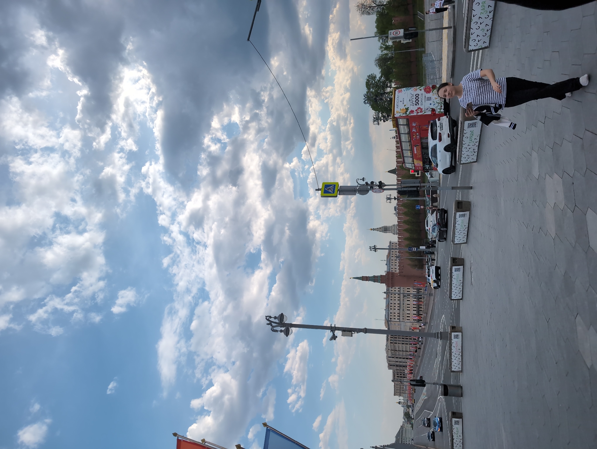 The image depicts a street scene in Moscow, Russia, likely near Red Square. A person wearing a striped shirt and dark pants is walking on a paved walkway. In the background is a red brick structure identifiable as part of the Moscow Kremlin. Various buildings line the street, and traffic is visible. A red double-decker tourist bus and several cars are present. Street lights, signs, and other urban infrastructure elements are also visible. The sky is filled with clouds.