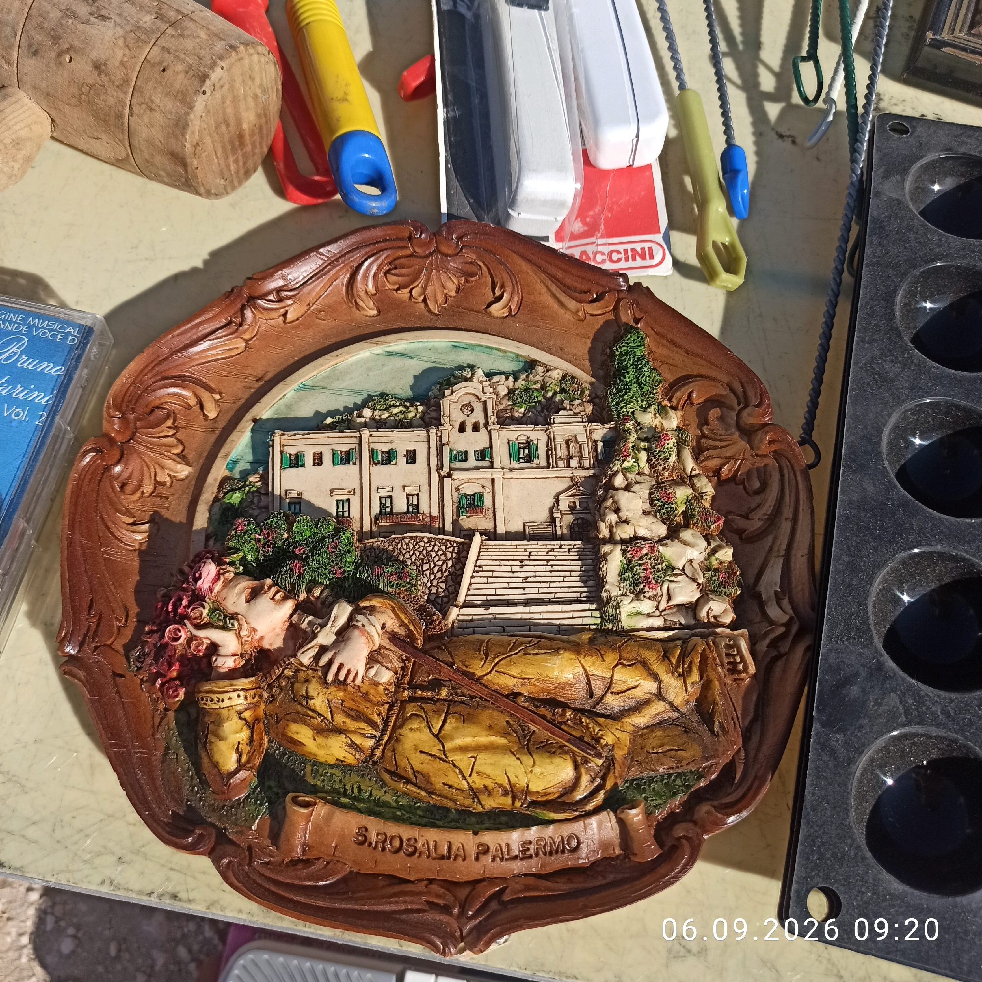A close-up, eye-level, medium shot captures a carved, oval-shaped wooden plaque depicting a scene of a woman lying amongst flowers with a building and stairs in the background. The plaque has an ornate, brown wooden frame. The woman is depicted with her eyes closed, holding a long, thin stick. The building appears to be white with green shutters. The plaque is resting on a light-colored, possibly marble or laminate, surface. To the left of the plaque is a clear plastic cassette tape case with blue and white text. Further left and slightly out of focus is a wooden mallet. In the upper part of the image, out of focus, are various colorful objects including a red and yellow toy with blue accents. To the right of the plaque is a black, multi-holed plastic object, possibly a mold or tray. The scene appears to be an outdoor market or yard sale, indicated by the variety of objects. The timestamp "06.09.2026 09:20" is superimposed on the bottom right of the image.