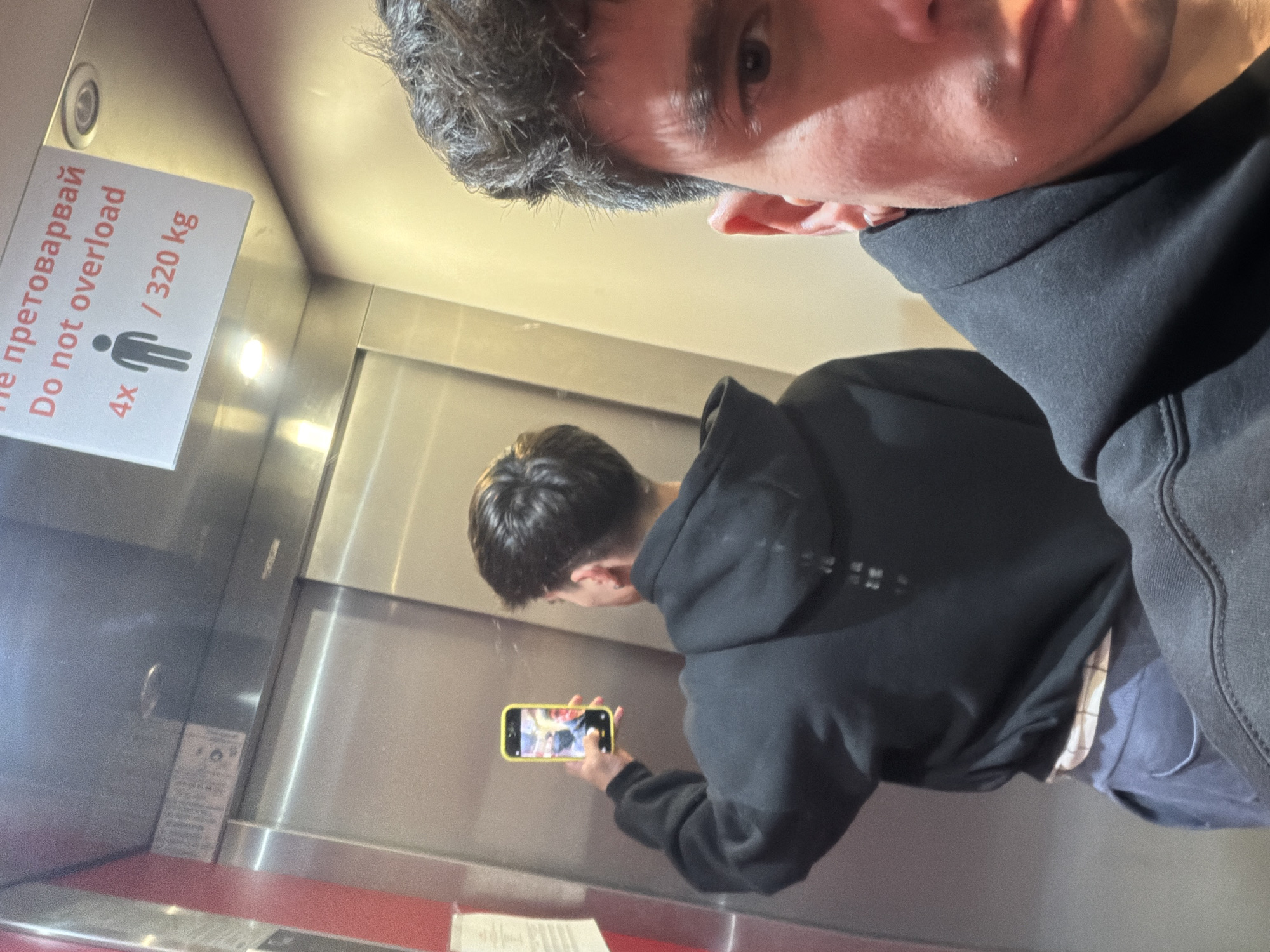 A selfie shows two males in an elevator. The elevator has a sign written in Bulgarian that says, "Не претоварвай" and "Do not overload", 4x, and /320 kg. One male, with dark hair, is in the foreground wearing a black hoodie. His face is partially visible. The other male, who appears to be younger, is wearing a black hoodie and holding a yellow smartphone, possibly taking a photo. The elevator interior is metal. Based on the Bulgarian text on the sign, the location is likely Sofia, Bulgaria.