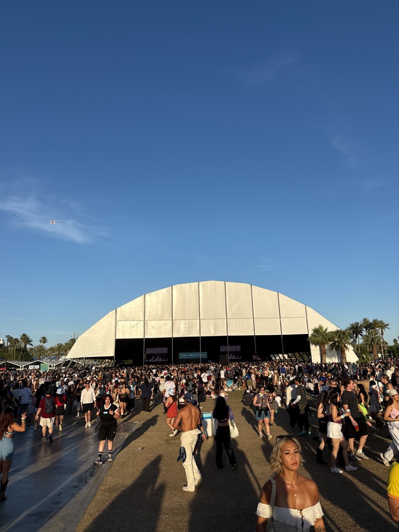 The image captures a vibrant outdoor scene, likely at the Coachella Valley Music and Arts Festival in Indio, California, United States. A large, white tent-like structure stands in the background, serving as a performance stage or enclosed area. A crowd of people fills the foreground, indicating a bustling festival atmosphere. Palm trees dot the landscape, contributing to the festival's characteristic desert setting. The clear blue sky suggests sunny weather, typical of the region.
