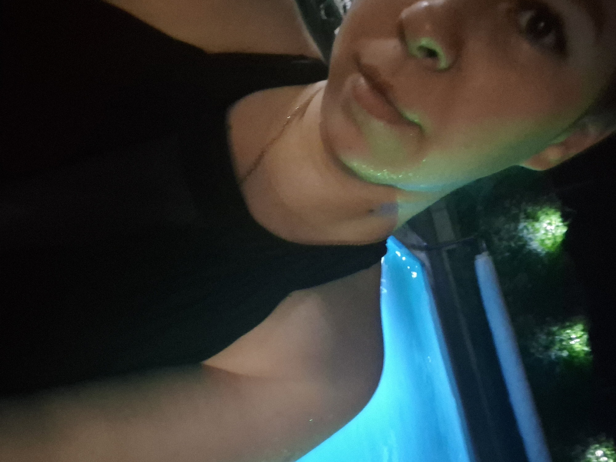 A person is at the edge of a pool at night. The pool water is lit with a bright blue light. Beyond the pool is a dark area with some bright spots of green, possibly plants illuminated by artificial light. The person is wearing a dark top and a thin gold necklace. The location is Chaikovsky, Russia.
