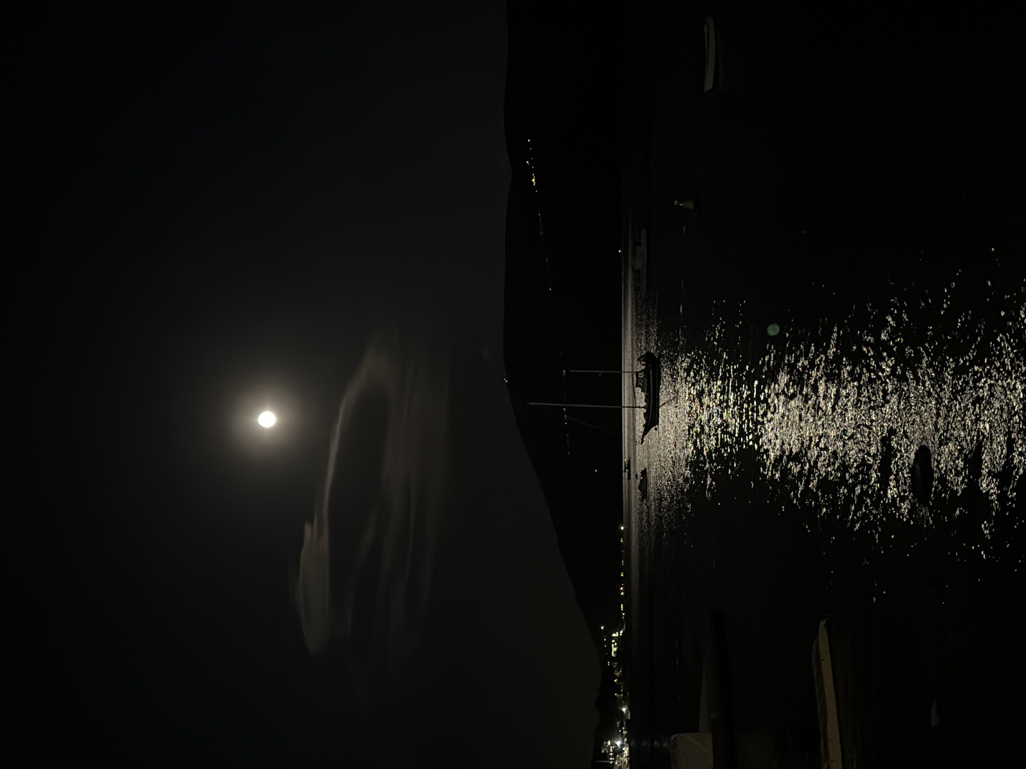 The image captures a peaceful nocturnal scene in Meljine, Montenegro, viewed in landscape orientation. The dark night sky fills the upper left portion of the frame, featuring a prominent, bright full or near-full moon. Faint, wispy clouds are subtly visible around the moon, adding a soft glow to the otherwise dark expanse.
A dark, indistinct landmass or elevated coastline stretches horizontally across the middle of the image, separating the sky from the water below. This landmass is mostly silhouetted, with a few tiny distant lights discernible along its base, suggesting a distant town or scattered buildings.
The lower half and right side of the image are dominated by a body of water, likely the sea or a bay, which reflects numerous artificial lights. These reflections appear as scattered, sparkling points and streaks of light, particularly dense and bright on the right side of the water surface. The shimmering effect suggests a gentle ripple on the water. A vague, dark silhouette that could be a small boat, pier, or part of a coastal structure is faintly visible near the water's edge on the far right, partially outlined by the reflected light.
The scene is devoid of explicit human activity. The overall impression is one of serene calm and natural beauty under moonlight, contrasted with the subtle glow of distant human habitation. No visible text is present in the image.