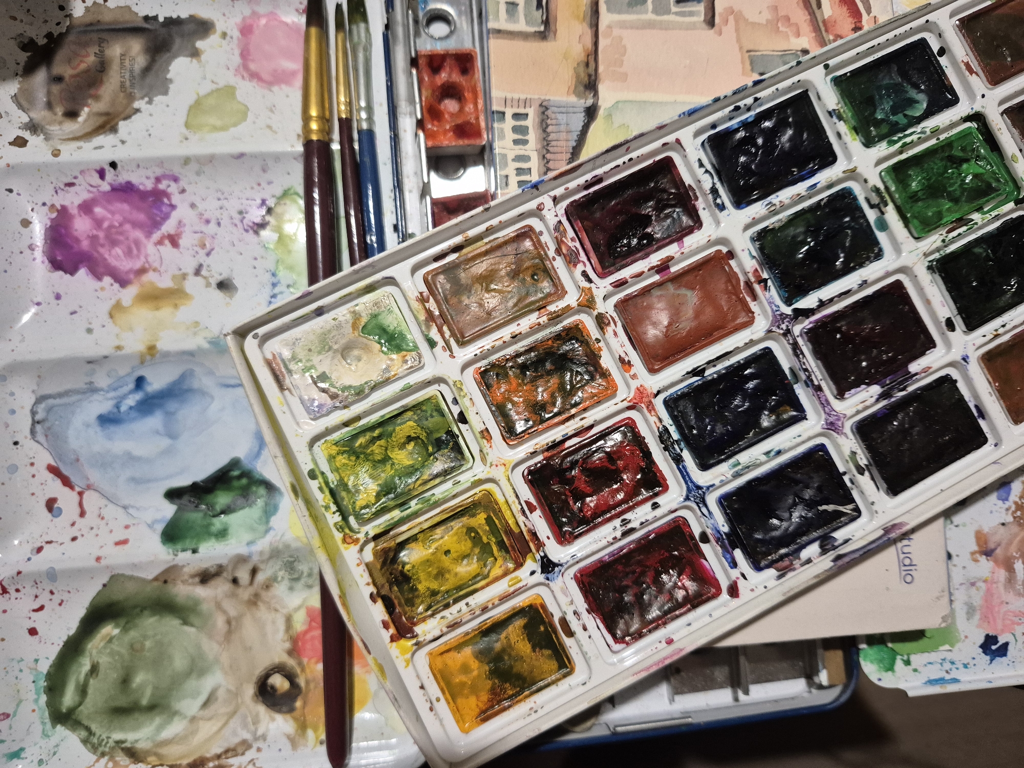 The image shows an overhead view of an artist's watercolor paint set. The paint set is a rectangular tray with multiple individual paint wells, most of which contain colorful, partially used watercolor paints. Brushes are visible resting on top of and beside the paint set. The tray itself is speckled with dried paint, indicating its use. In the background, another artist's palette with splotches of paint and a partial view of a watercolor painting of buildings are visible. The overall scene suggests an art studio or creative workspace. The location is not discernible from the image.