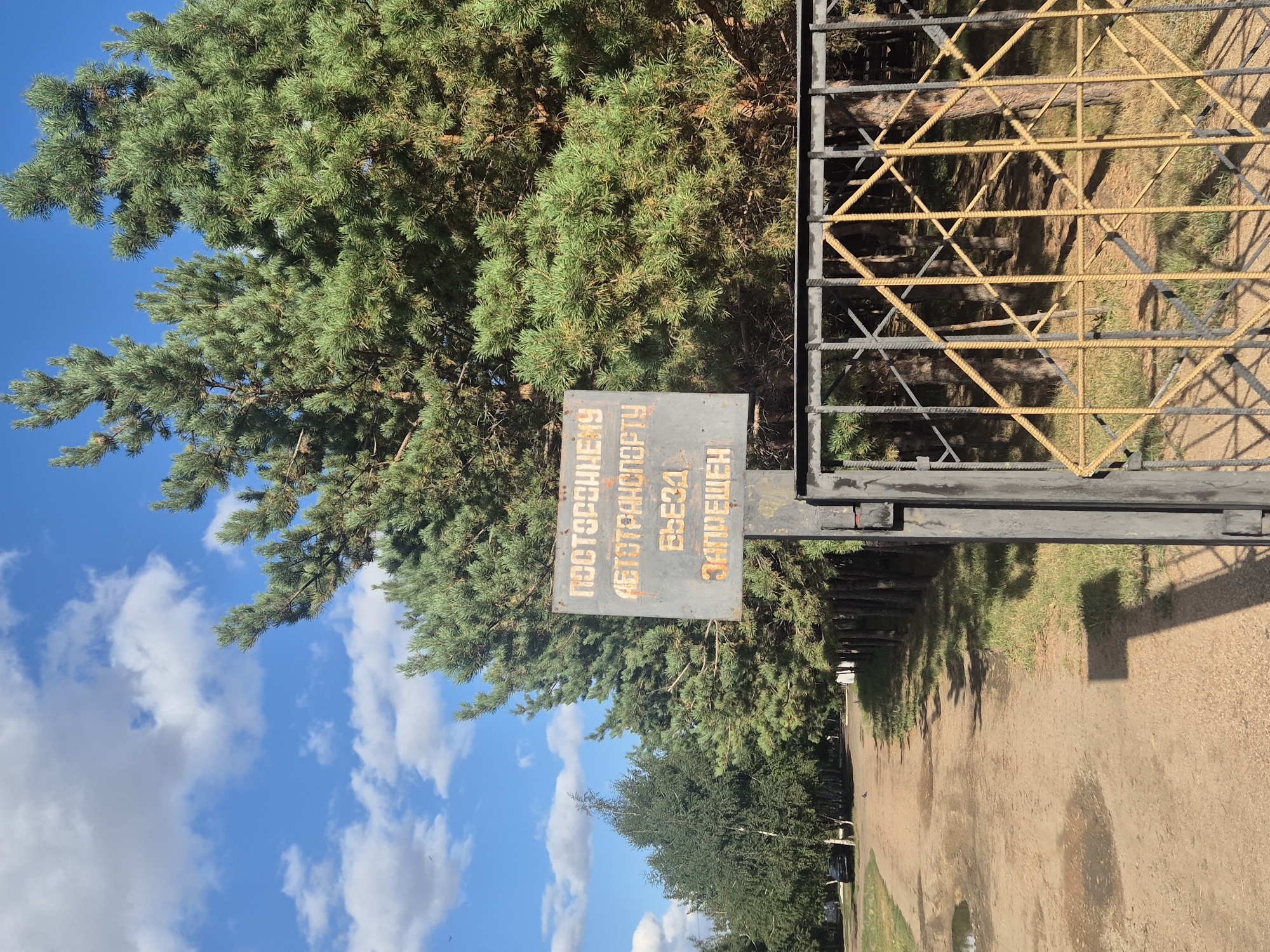 The image shows a sign in front of a metal gate, with trees and a dirt road in the background. The sign is weathered and has text in Russian that reads "ПОСТОРОННЕМУ АВТОТРАНСПОРТУ ВЪЕЗД ЗАПРЕЩЕН," which translates to "Entry forbidden for foreign vehicles." The gate is made of black metal bars with yellow diagonal reinforcements in a diamond pattern. Behind the gate, there are tall pine trees. To the left of the gate, a dirt road curves into the distance, lined with trees on the right and a row of trees on the left. A black car is parked on the side of the road in the distance, on the left. The sky is blue with scattered white clouds. The overall scene suggests a restricted area or private property. This location is in Kokshetau, Kazakhstan.