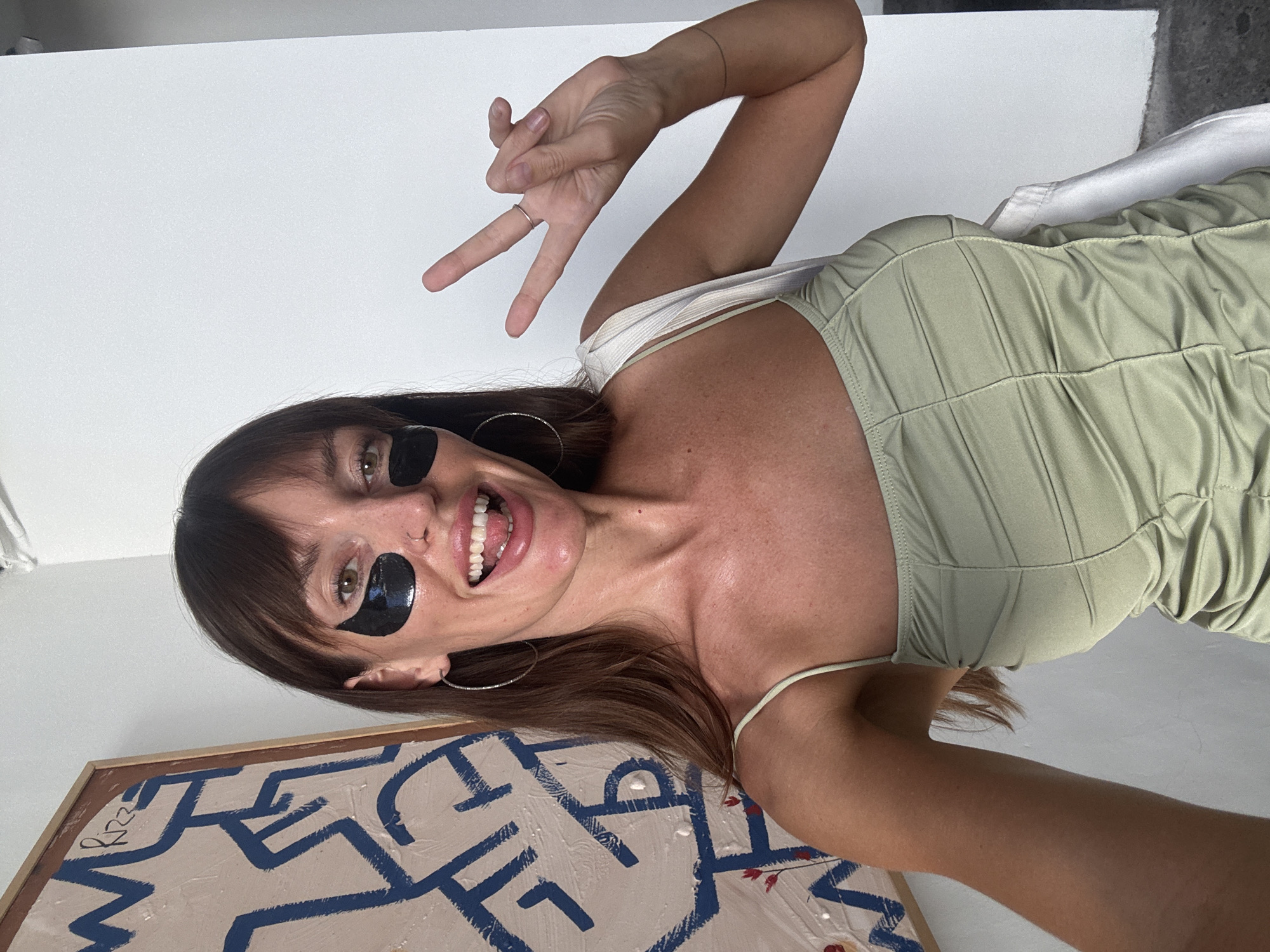 A woman is facing the camera, with her head tilted to the left, wearing a light green sleeveless top and black under-eye patches. Her brown hair has bangs. She is flashing a peace sign with her right hand. A painting with a blue abstract design on a beige background is located below her and slightly to the left. The background is a plain white wall. This image was likely taken indoors. It is not possible to determine whether this photo was taken in North Kuta, Indonesia.