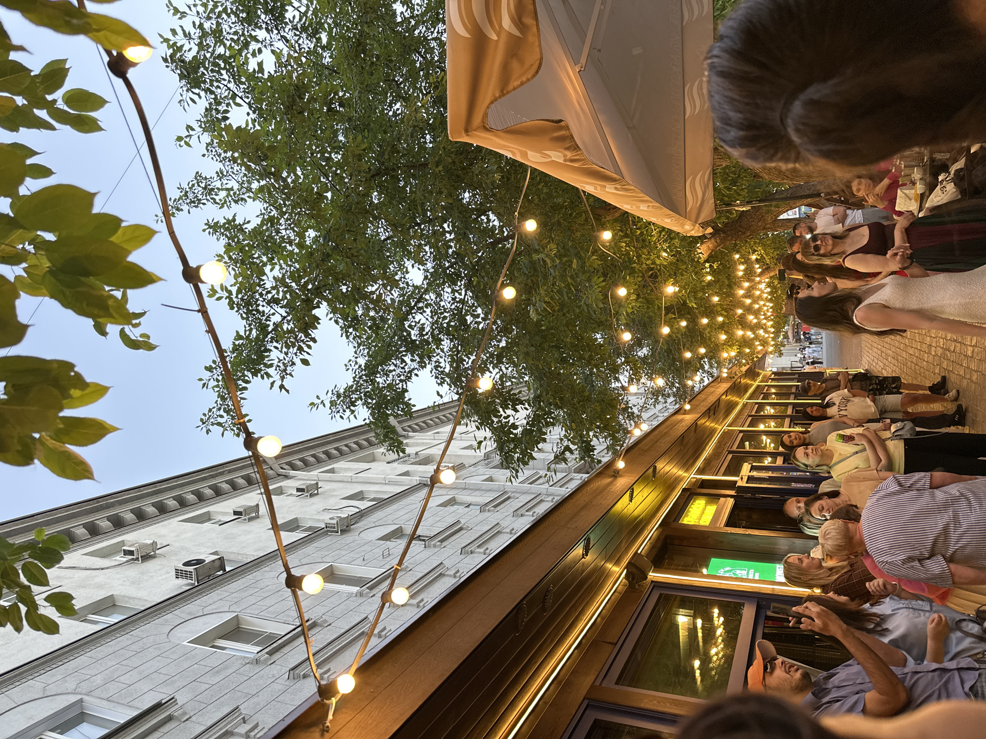 The image depicts an outdoor seating area of a restaurant or cafe on a street in Novosibirsk, Russia. String lights illuminate the scene, creating a warm ambiance. On the left, a building with multiple windows and air conditioning units lines the street, with awnings and lights extending from its facade. In the foreground and midground, several people are gathered, some standing and talking, while others appear to be waiting or dining. The people are dressed casually, suggesting a relaxed atmosphere. To the right, a large umbrella or awning provides shade, and trees with lush green foliage contribute to the natural setting. The overall impression is a lively urban outdoor space, likely enjoyed during an evening or late afternoon.