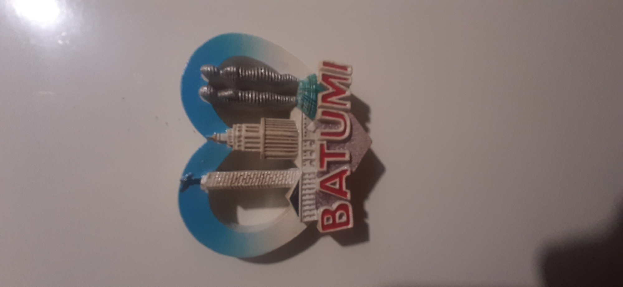The image is a close-up, slightly angled shot of a magnet shaped like a heart with a stylized representation of Batumi, Georgia, within it. The magnet features several iconic landmarks of Batumi: a tall, slender tower topped with a figure, a cylindrical building with multiple tiers, and two intertwined metallic figures. The word "BATUMI" is prominently displayed in red, raised lettering against a light-colored background at the bottom of the heart shape. The background of the magnet is a gradient of blue, transitioning from a lighter shade at the top to a darker shade at the bottom. The magnet is affixed to a plain, light-colored surface, likely a refrigerator. There are no people or animals visible in the image. The lighting suggests an indoor environment, with some glare from a light source visible in the upper right corner. The image does not provide any information related to Yessentukskaya, Russia.