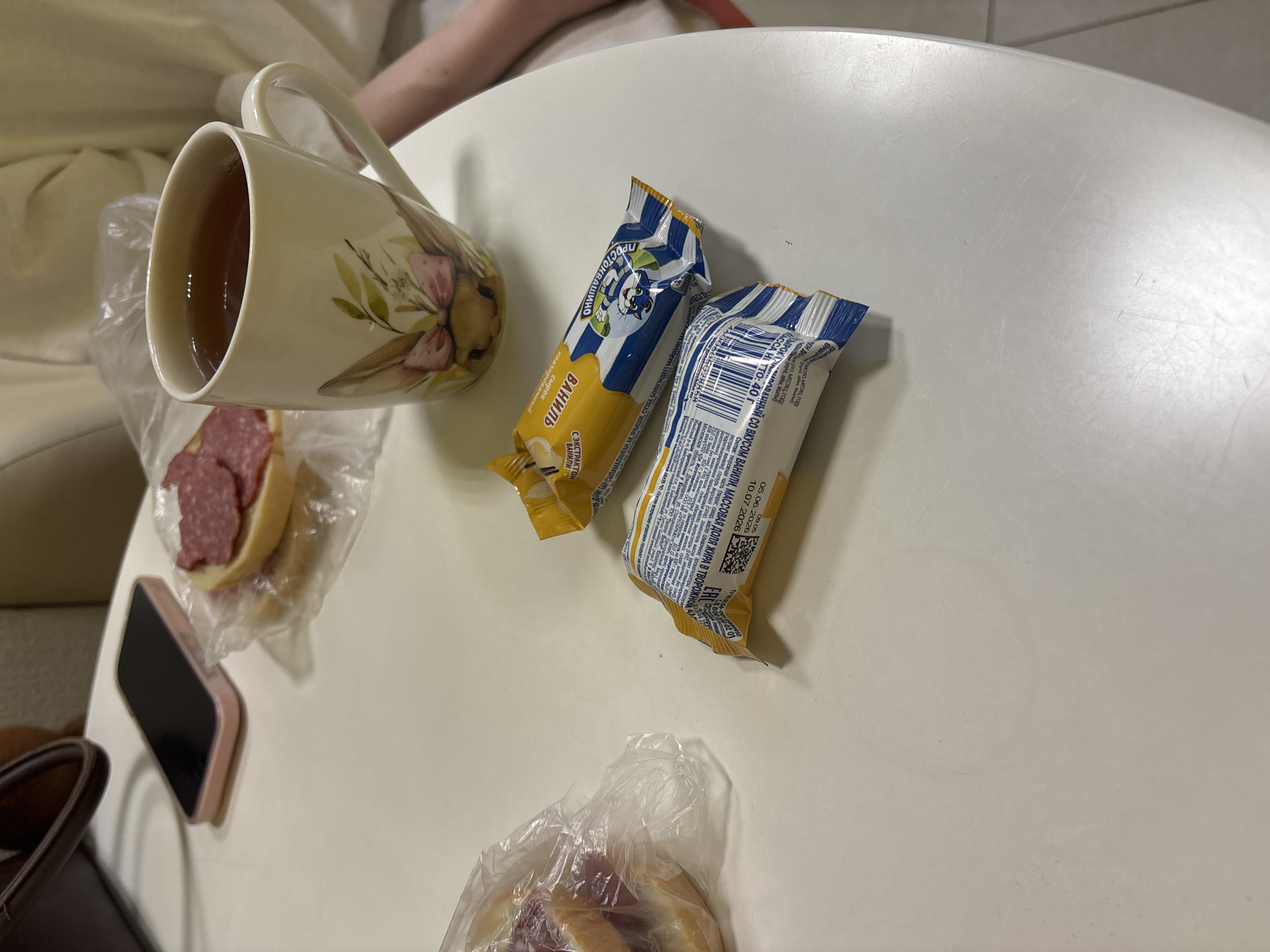 On a round, white table, an overhead shot captures a casual snack. In the foreground, two foil-wrapped snack bars, one with blue and white stripes and a cartoon character, and another with a yellow and blue wrapper, lie side-by-side. To the left, a sandwich with sliced salami on a bun is partially visible within a clear plastic bag. Behind it, a pink smartphone rests on the table. A ceramic mug, adorned with a watercolor painting of a rabbit and flowers, is filled with a dark liquid, likely tea, and sits to the right of the sandwich. In the background, blurred, light-colored fabric, possibly clothing or a tablecloth, creates a soft backdrop. The scene suggests a moment of informal dining or a break. This image does not provide information that would allow for location determination in Sinyavino, Russia, or any other specific geographic location.