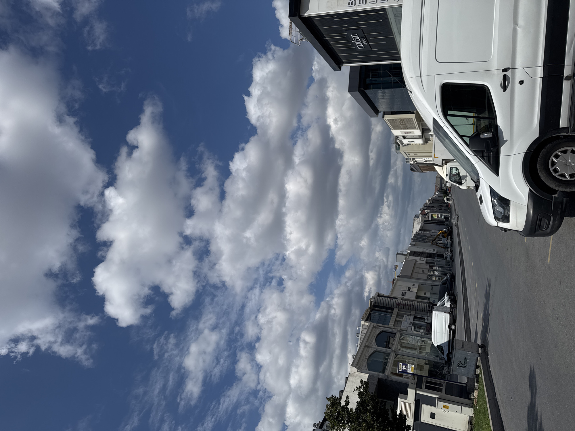 The image shows a street scene in Güngören, Turkey, under a partly cloudy blue sky. A white van is parked on the right side of the street, with its front visible. In the background, several other vans and vehicles are parked along the roadside. Buildings with modern architecture line the street, some featuring large windows and signage. A tree is visible on the left side of the image. The overall scene depicts a commercial area on a bright day.
