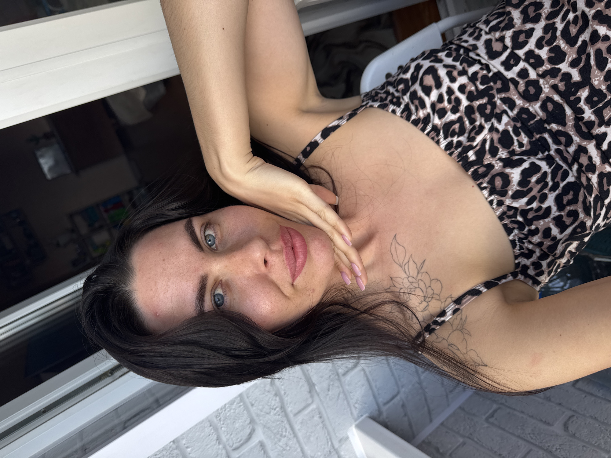 A young woman with long, dark hair and blue eyes takes a selfie. She has light skin with a few blemishes and is wearing a leopard-print dress. Her right hand is resting on her chin and her arm is raised, with her hand touching her face. A tattoo of flowers is visible on her chest and shoulder. She is standing on a balcony with a white brick wall to her left and a window to her right. The background shows an interior room with furniture. The image was taken in Chaikovsky, Russia.
