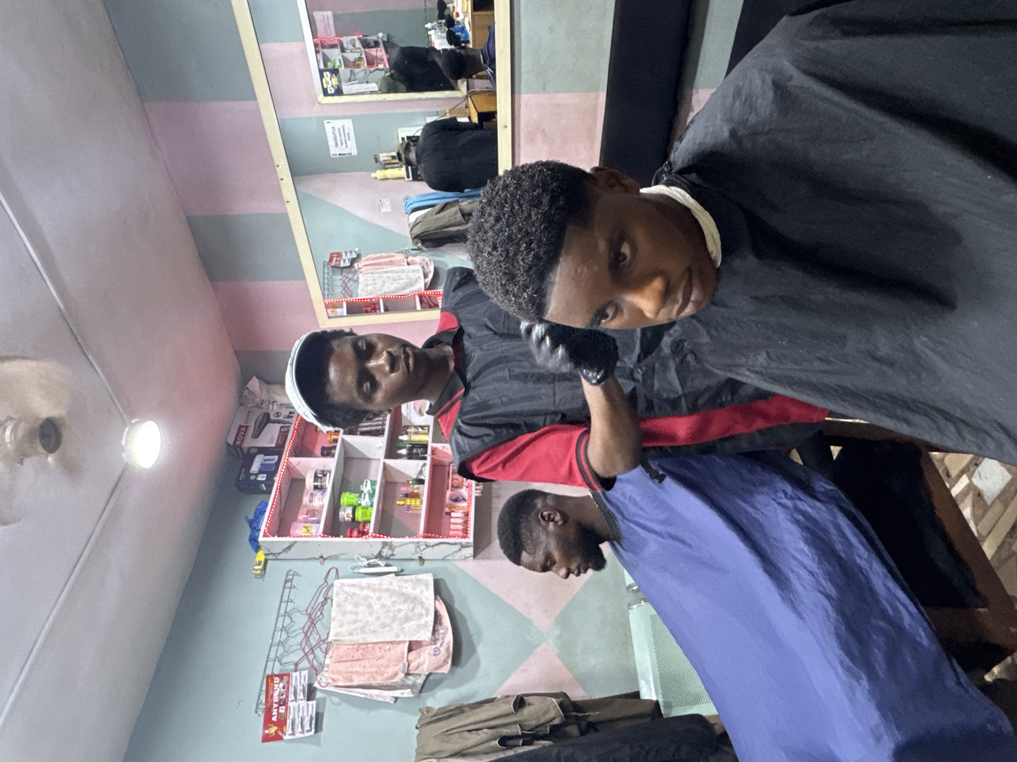 The image depicts a barber shop scene in Nkpor, Nigeria. A young man is seated, draped in a black cape, receiving a haircut. The barber, wearing a white cap and a red and black jacket, is focused on the haircut. Another individual, covered in a blue cape, is visible in the background. The shop's interior has pastel-colored striped walls and a mirror reflecting additional patrons. A shelf unit with various hair products and a clothes rack with towels are also visible.