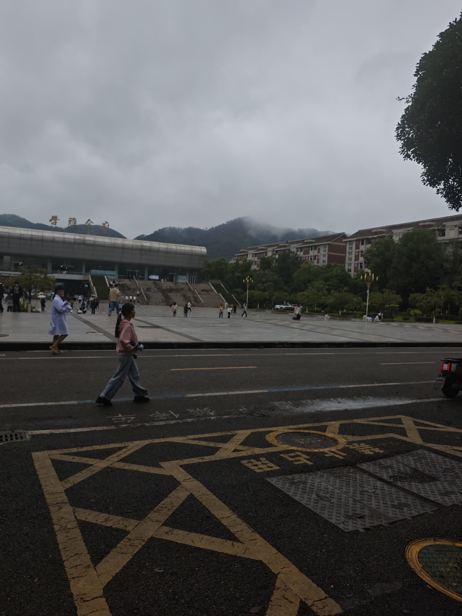 In the Chongqing High-tech Zone, China, on a cloudy day, a young girl in a pink hoodie and jeans walks across the street, looking to her left. She is in the foreground of the image, with a large, modern building with a wide stairway and signage in Chinese characters behind her. The building is set back from the street and is surrounded by a large, paved plaza where other pedestrians are present. Beyond the plaza, there are apartment buildings with red roofs and lush green trees. Mountains shrouded in mist are visible in the background. The street in the foreground has yellow road markings, including a large 'X' shape and what appear to be pedestrian crossing indicators. A section of a black vehicle is visible on the right side of the image.