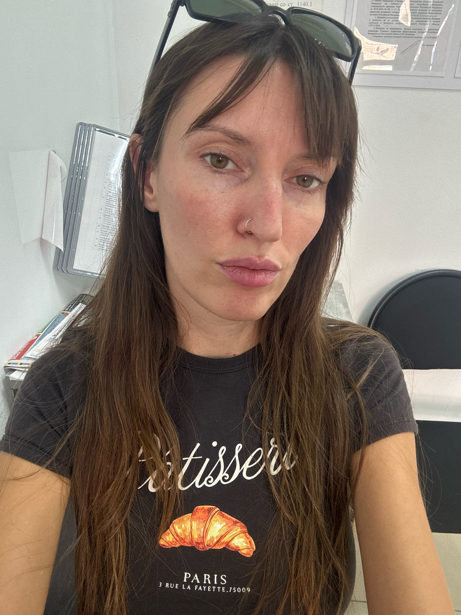 This is a close-up selfie of a woman with long, dark brown hair and bangs, wearing sunglasses on top of her head. She has a nose ring and is looking directly at the camera with a neutral expression. She is wearing a dark t-shirt with a graphic print of a croissant and text that reads "tisser PARIS 3 RUE LA FAYETTE, 75009".
The background suggests an indoor setting, possibly an office or waiting room, with files and papers visible on a shelf to her left, and a black, semi-circular object to her right. The lighting appears to be natural and consistent, suggesting it could be daytime. There is no clear indication of the weather or a specific event taking place. The text on the t-shirt points to a Parisian theme, but the location context provided is Bataysk, Russia.
