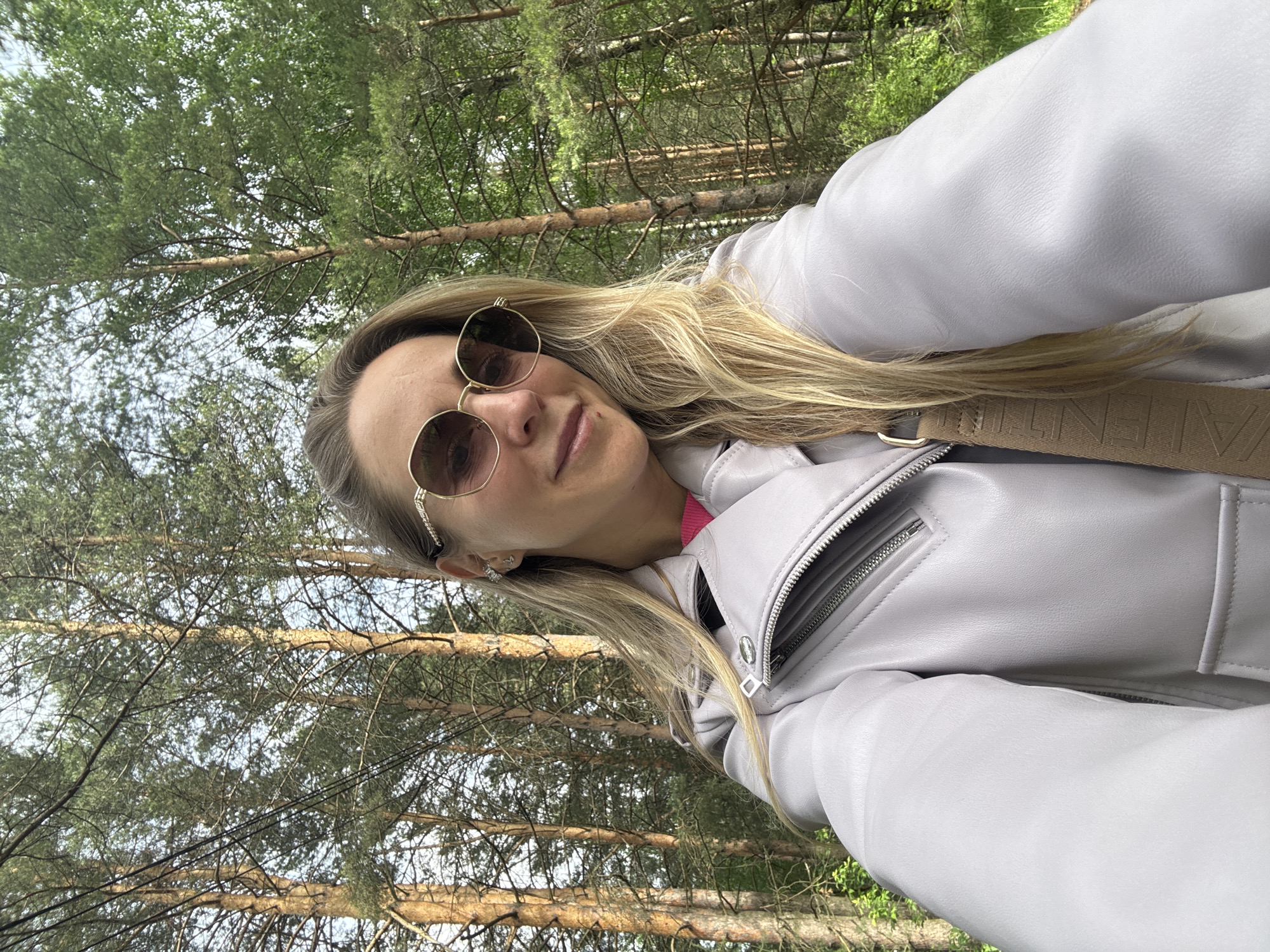 A blonde woman is captured in a selfie taken outdoors in a forest, with the image rotated 90 degrees counter-clockwise. She is positioned in the foreground, looking directly towards the camera with a calm, slightly smiling expression. She wears stylish round, gold-rimmed sunglasses with tinted lenses, which partially obscure her eyes. Her long, blonde hair flows over her shoulders and jacket.
She is dressed in a light gray or lavender faux leather jacket, featuring prominent silver zippers on the front and pockets. A bright pink top or shirt is visible underneath her jacket. A beige strap, likely from a shoulder bag, is visible across her chest, with the brand name "VALENTINI" clearly embossed on it. A small, sparkling earring adorns her right ear.
The background is dominated by a dense coniferous forest, filled with numerous tall pine trees. Their green needles and brown trunks create a natural, serene setting. Patches of bright sky are visible through the tree canopy, indicating it is daytime. The overall lighting is bright and even, suggesting pleasant weather for an outdoor activity like a walk or hike. The scene is consistent with the natural landscapes found in Chaikovsky, Russia.