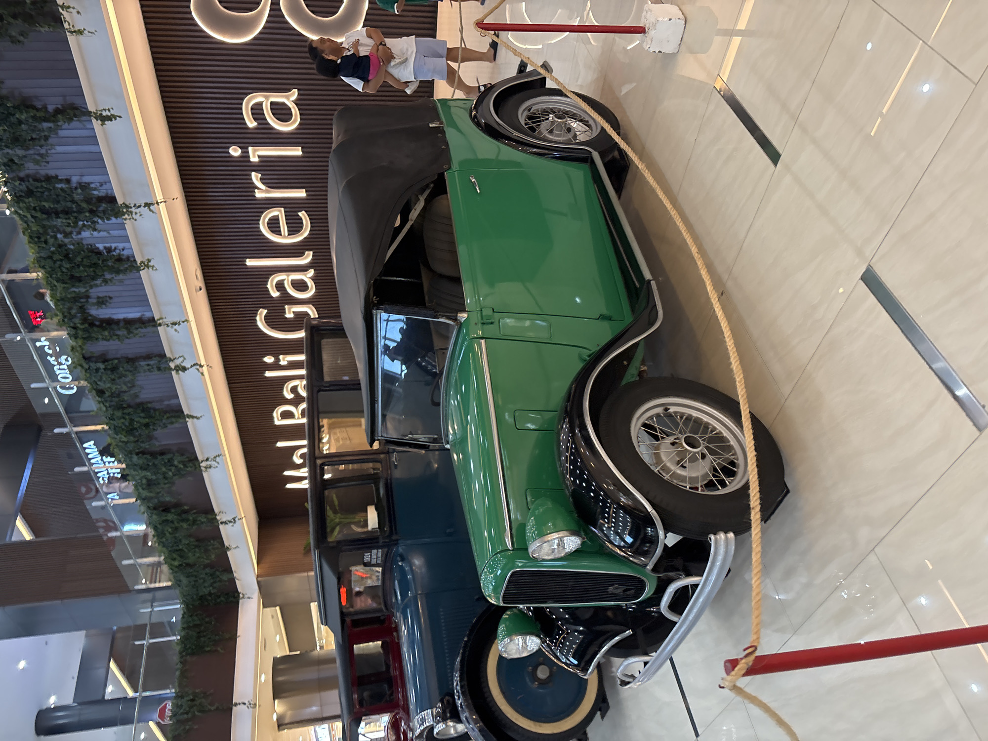 The image shows a display of vintage cars at the Mal Bali Galeria shopping mall in Kuta, Indonesia. A green convertible is prominently displayed, roped off, in the foreground. Behind it, other vintage cars are lined up, partially visible in red and blue. Shoppers are walking past the display. The scene is indoors with bright overhead lighting and a polished tile floor.