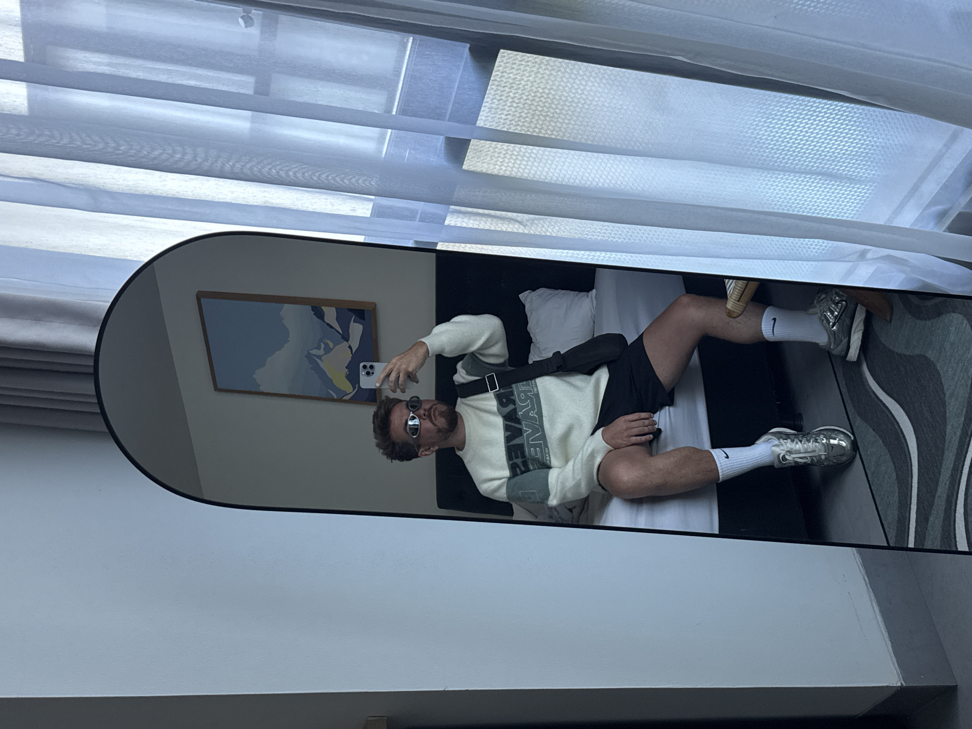 The image shows a man reflected in a full-length mirror. He is lying on a bed, wearing sunglasses, a light-colored sweater with dark text, black shorts, white socks with a Nike logo, and silver sneakers. A black shoulder bag is slung across his body. He is holding a smartphone and appears to be taking a selfie. Behind him are white pillows, a headboard, and a framed abstract painting on the wall. The room has sheer curtains and a gray rug with a wavy pattern. The location is North Kuta, Indonesia.