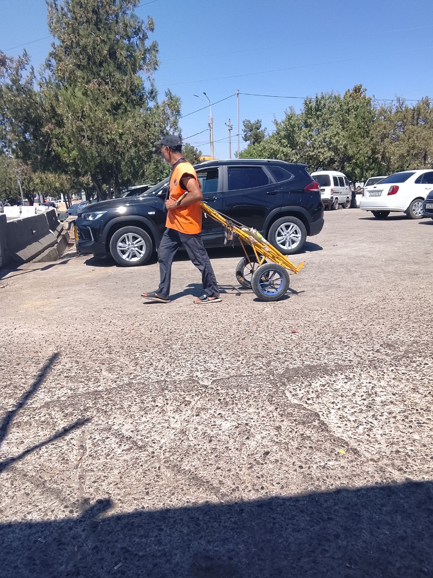 A man walks a yellow wheeled cart across a paved lot in Tashkent, Uzbekistan. The man wears an orange vest and dark pants. The cart has two large wheels and appears to be made of metal. In the background, a black SUV, a white van, and a white sedan are parked. Trees line the background of the scene under a clear blue sky. The ground is cracked and weathered asphalt.