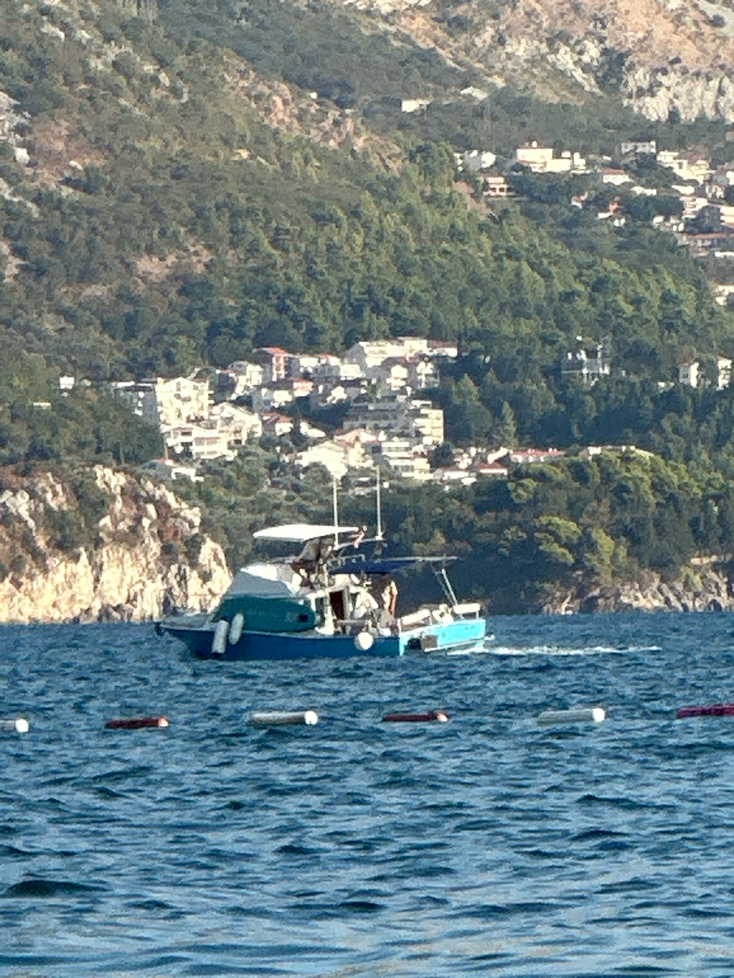 A motorboat with a white canopy is traveling on the water. The boat is blue and green with white fenders. There are at least two people visible on the boat. In the background, there is a hillside covered with green trees and dotted with white buildings. There are also rocky cliffs visible. The water is a deep blue with ripples. Several buoys are floating in the water in the foreground. The scene depicts a coastal area near Boreti, Montenegro, likely a resort town given the buildings on the hillside.