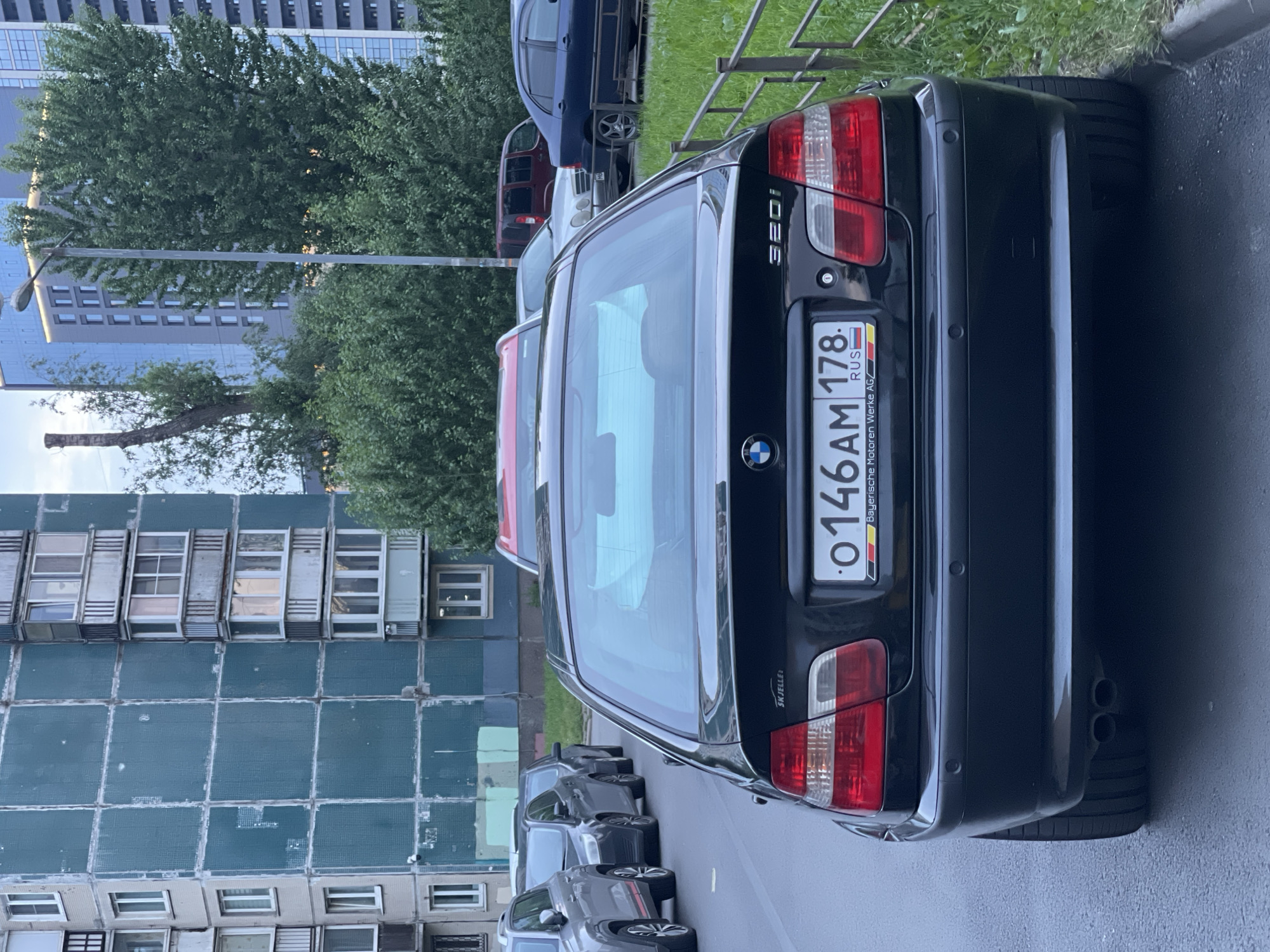 A black 2002 BMW 320i sedan is parked on an asphalt street. The car's license plate reads "0 146 AM 178". The car is positioned parallel to a curb, with grass and a metal fence to its right. In the background, there are several other vehicles, including a silver SUV, a dark sedan, and a red minivan. Further in the background, there are apartment buildings and trees. The sky is overcast.