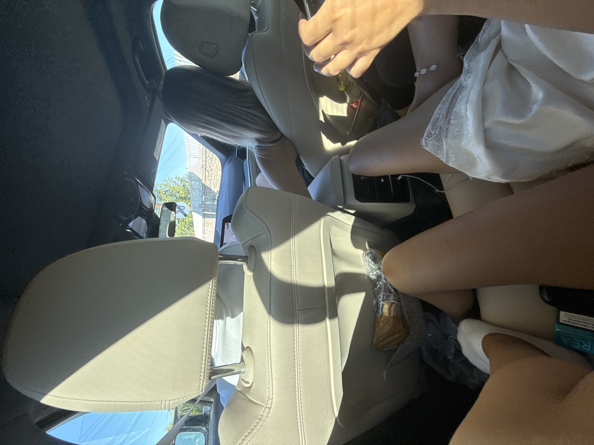 The image shows the interior of a car, viewed from the back seat looking forward. Two women are seated in the back. The woman on the left has her legs propped up and is wearing a white sock. The woman on the right has her legs extended and is wearing a white lace garment and a seashell anklet. A hand, presumably belonging to another passenger, is visible in the foreground on the right. There are items in the seatback pockets, including a wrapped snack and a bottle. Outside the car, a white fence and trees are visible under a clear sky. A speed limit sign displaying "30" is mounted on a post outside. The scene does not provide any specific information to place it in Simila, Romania.