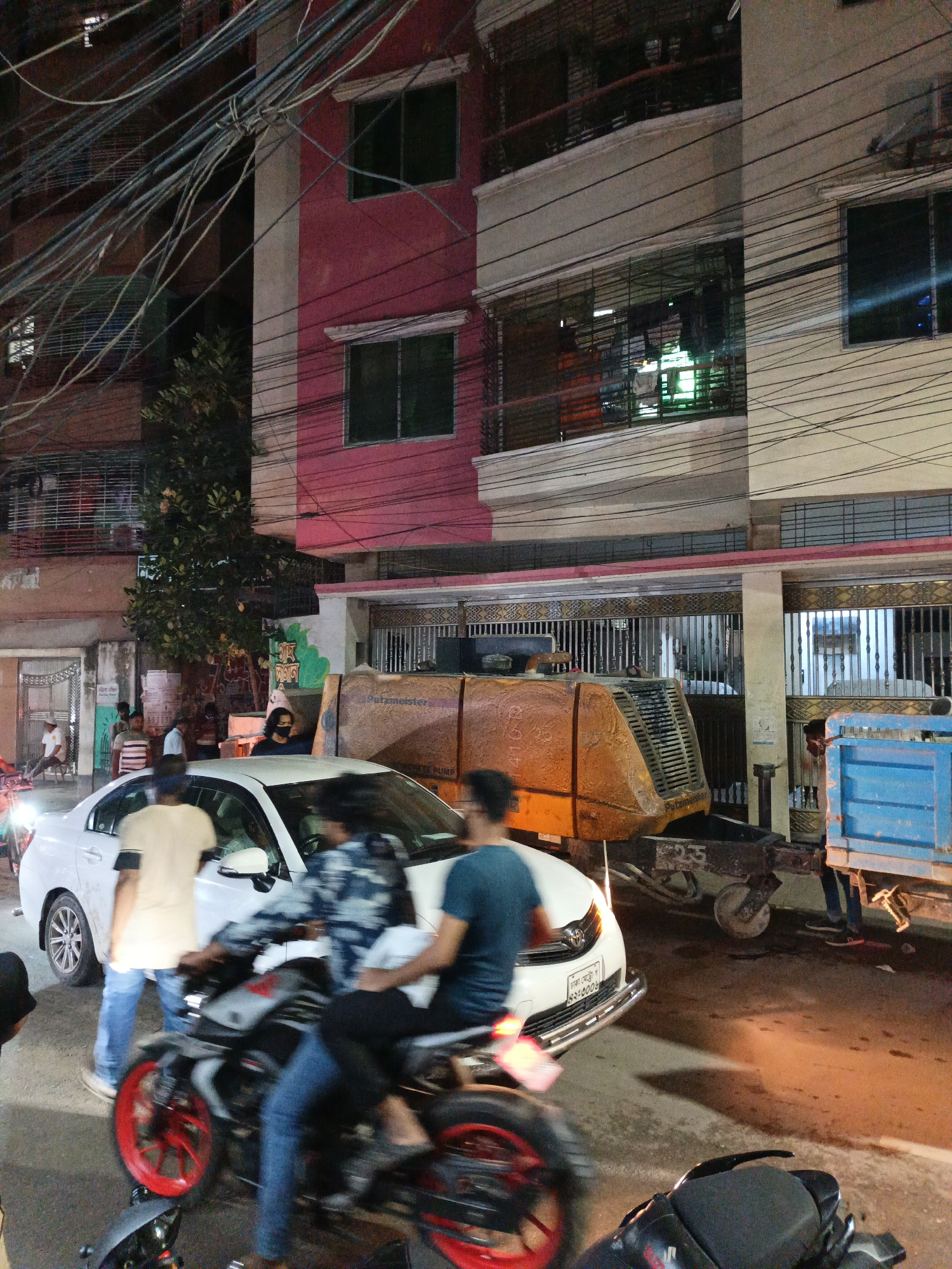 The image captures a bustling urban street scene at night, likely in Dhaka, Bangladesh, as evidenced by the Bengali script "ঢাকা মেট্রো ঘ ৪১-৪০০৯" (Dhaka Metro Gha 41-4009) on a white car's license plate. The scene is illuminated by artificial light, creating a vivid, high-contrast atmosphere.
In the upper portion of the frame, a dense and tangled network of overhead electrical or communication wires spans across the street, connecting various buildings and utility poles. The background features multi-story buildings, some with prominent pink/red facades and others in lighter tones, displaying windows and balconies with metal railings.
The midground and foreground are occupied by people and vehicles. A white sedan is prominent, with an individual in a beige shirt standing near its front passenger side door. Another person, wearing a black face mask, stands further back near the car. Two motorcycles are captured in motion in the foreground, their riders and parts of the bikes (including one with bright red rims) significantly blurred, indicating movement.
To the right, a large, yellow-orange industrial machine, identified by visible text as a "Putzmeister CONCRETE PUMP" with model numbers such as "410 D" and "K-235", is parked or being moved. It appears to be attached to a trailer. Adjacent to this machinery is the rear section of a blue truck or trailer with a cargo bed. The overall scene conveys a lively, active urban environment after dark.