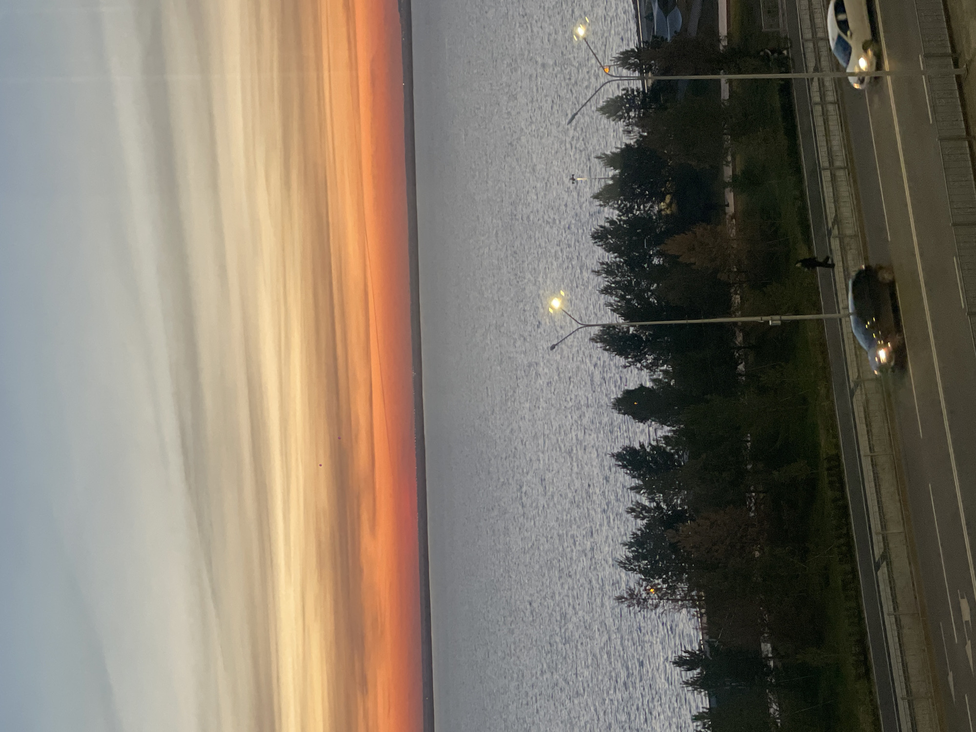 A horizontally-oriented photograph captures a view of a city beside a body of water under a dynamic sky. The left side of the image features a sunset-colored sky in shades of orange and gray. Adjacent to the sky is a large body of water, possibly a lake or bay. Along the water's edge, a line of trees is visible, interspersed with streetlights. Further to the right, a road runs parallel to the water, with at least two vehicles, one white and one dark, traveling on it. A lone pedestrian is present on the sidewalk between the trees and the road. Based on the landscape and buildings in the image, it appears to be a view of Kokshetau, Kazakhstan, specifically the area where the city meets a large body of water like Lake Kopa.