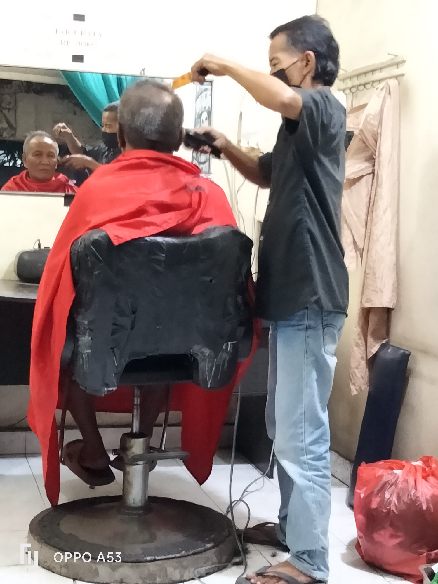 In a humble barbershop in Denpasar, Indonesia, a barber, wearing a dark t-shirt and a face mask, is meticulously cutting the hair of an elderly man. The customer is seated in a black, slightly worn barber chair, draped in a vibrant red cape. The reflection in the mirror behind them reveals a closer view of the barber's hands at work, and the customer's face, which shows a calm expression. The barbershop appears simple, with a white wall, a mirror, and some shelves. A light-colored garment hangs on a rack in the background, and a large red plastic bag lies on the floor, suggesting it might contain discarded hair clippings. The overall atmosphere is one of quiet efficiency, capturing a common scene of everyday life in a local Indonesian community.