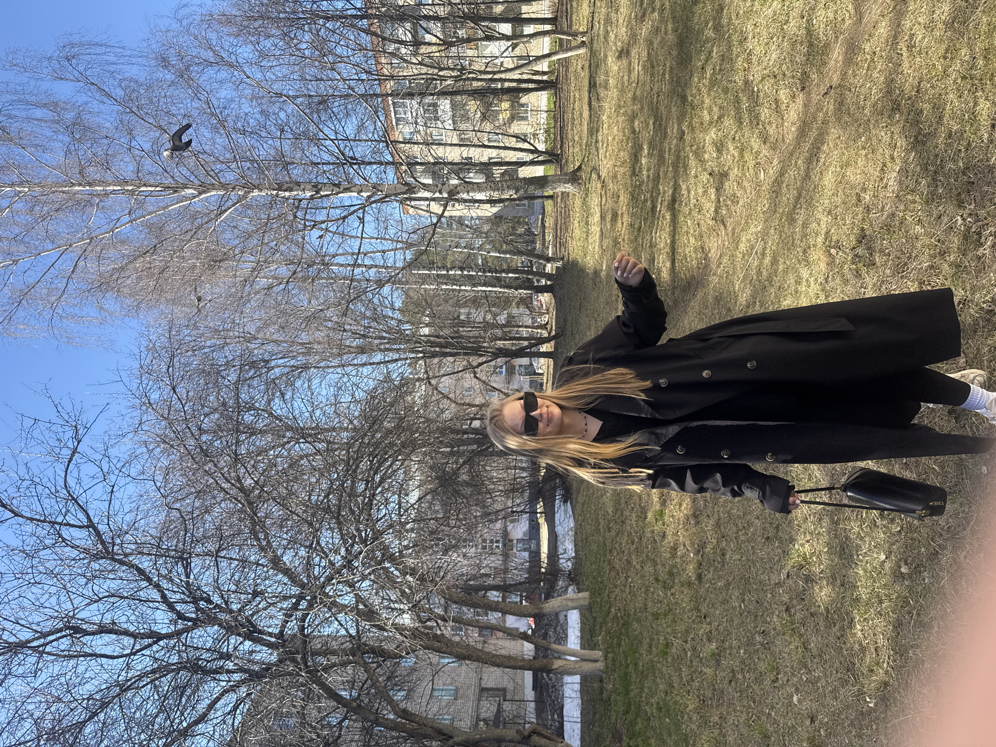 A medium shot, landscape oriented, shows a woman wearing a long black trench coat and sunglasses, carrying a black bag, standing on a grassy lawn, likely in a park in Chaikovsky, Russia.
The woman is blonde and looks toward the camera. Behind her are bare deciduous trees against a clear blue sky, with a dark bird flying in the upper left. Beyond the trees is a multi-story apartment building. The grass is dry and brown, indicating it is early spring or late fall. A river is visible on the middle-left side of the image.