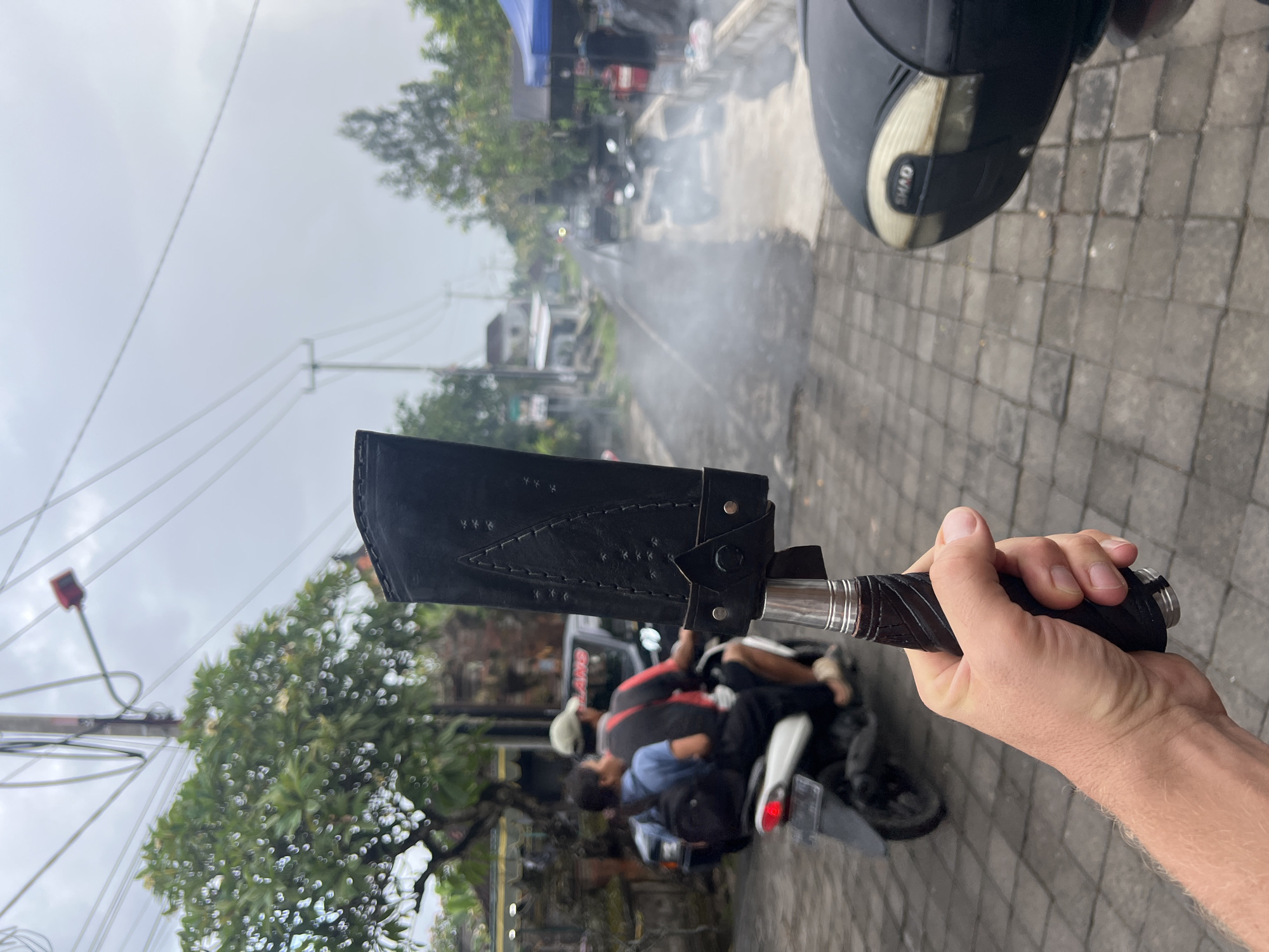 A person is holding a large knife in a black sheath while seated on a motor scooter in Sukawati, Indonesia. The knife is held upright in the right hand of a light-skinned individual, with the handle wrapped in black material. The sheath is also black and appears to be made of leather. A motor scooter with a black storage compartment is visible next to the person's hand. The background shows a street scene with several motorbikes parked. People are sitting on the motorbikes. Trees and buildings line the street, and overhead power lines are visible. The weather is overcast. There appears to be smoke or steam rising from the street in the background. The ground is paved with gray stones.