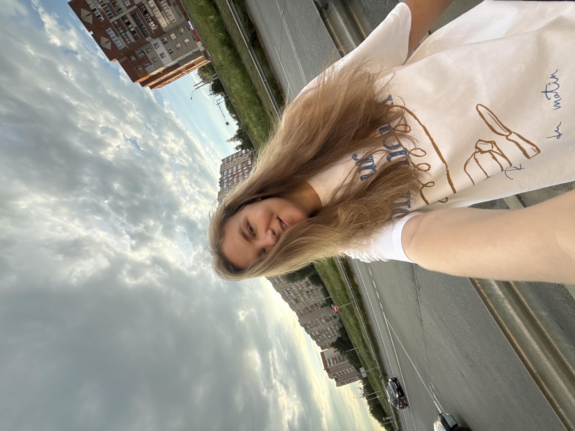 The image is a wide-angle, selfie-style shot taken outdoors, possibly in a city. A young woman with long blonde hair is the primary subject, positioned in the foreground and slightly to the right. She is wearing a white t-shirt with a graphic design. Her face is partially obscured by her hair, but she appears to be smiling.
In the background, there are several multi-story apartment buildings, characteristic of Soviet-era architecture, with balconies and numerous windows. These buildings are aligned along a road that curves to the left. The road is asphalt, with lane markings visible, and appears to be a busy thoroughfare, as at least two cars are present, one of which is a black sedan. A metal barrier separates the road from a grassy embankment. The sky is overcast with dramatic clouds, with patches of blue sky visible, suggesting either dawn or dusk.
Based on the architectural style of the buildings, the presence of specific road signage (a red circular sign with a white bar, indicating no entry), and the overall urban landscape, this image is likely from the city of Vladimir, Russia. Specifically, the composition, including the long apartment blocks and the elevated road, is consistent with residential areas on the outskirts of Vladimir.