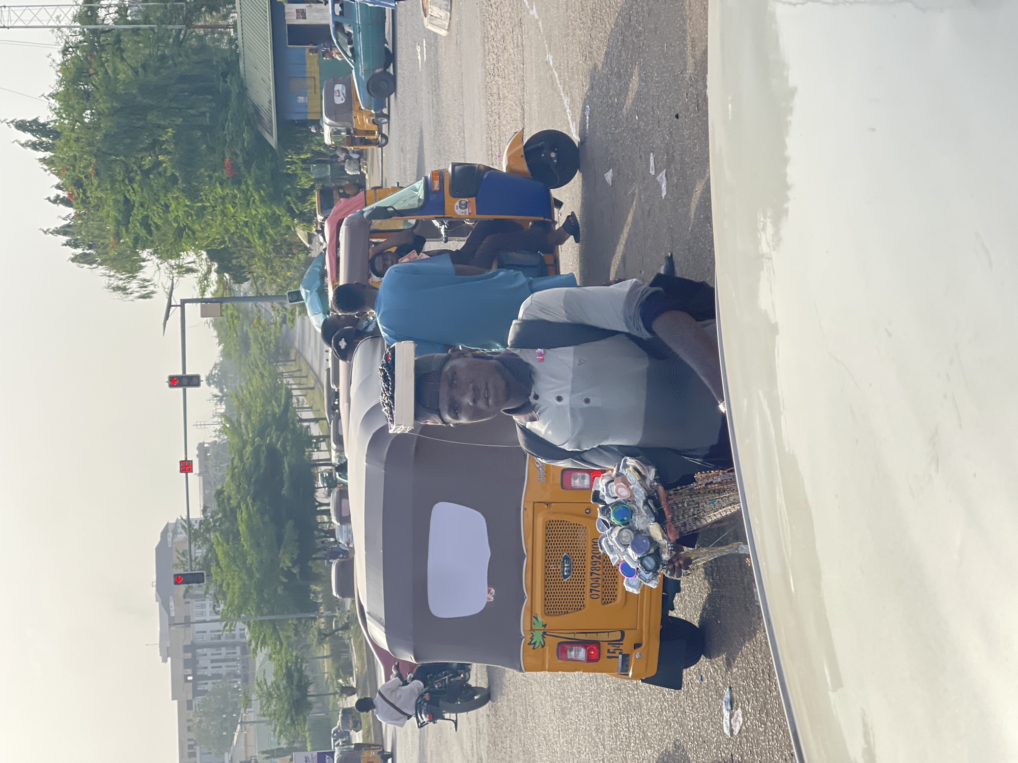 The image depicts a vibrant street scene in Abuja, Nigeria, during bright daylight hours, possibly late morning or early afternoon, with a slight atmospheric haze. The perspective is from within or beside a white vehicle, partially visible on the right.
The central focus is a line of auto-rickshaws, locally known as Keke Napep, stopped or moving slowly on a paved road, likely due to traffic. A man, appearing to be a street vendor, stands beside a prominent yellow and brown Keke Napep. He is wearing a light grey and white patterned shirt with a dark cap and looks directly at the camera. He holds a multi-tiered display of small items, including numerous colorful watches and various accessories, indicating he is selling goods to commuters.
Behind him, other people are visible in the queued traffic, some seated in other Keke Napeps. Further down the road, another blue and yellow Keke Napep is parked.
The background shows green trees lining the street, suggesting an urban boulevard, with buildings faintly visible through the haze in the distance. Traffic lights are mounted on poles overhead, with one displaying a red signal and a digital countdown showing "38". The overall impression is a bustling urban environment characterized by everyday street commerce and vehicular traffic.
Visible text includes a phone number "07047892000" and the number "154" on the rear of the yellow Keke Napep, and "HB" with a number "8" on the side of another Keke Napep further back.