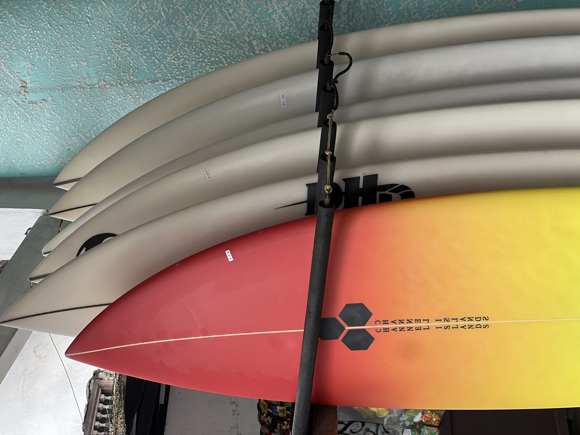 A close-up, eye-level shot shows a row of surfboards leaning against a textured, light blue wall. The surfboards are arranged vertically, with their tails facing upwards and their noses downwards. The surfboard on the left has a gradient of red and yellow, fading to orange at the bottom. It features black hexagonal shapes and vertical text that reads "CHANNEL ISLANDS". The other surfboards are lighter in color, appearing to be white or cream. A black padded rack, attached to the wall, supports the surfboards. The background reveals a glimpse of a building with a balcony and an ornate railing, suggesting an outdoor setting in an urban environment, possibly Kuta, Indonesia, known for its surf culture. The overall lighting is diffused, casting soft shadows.