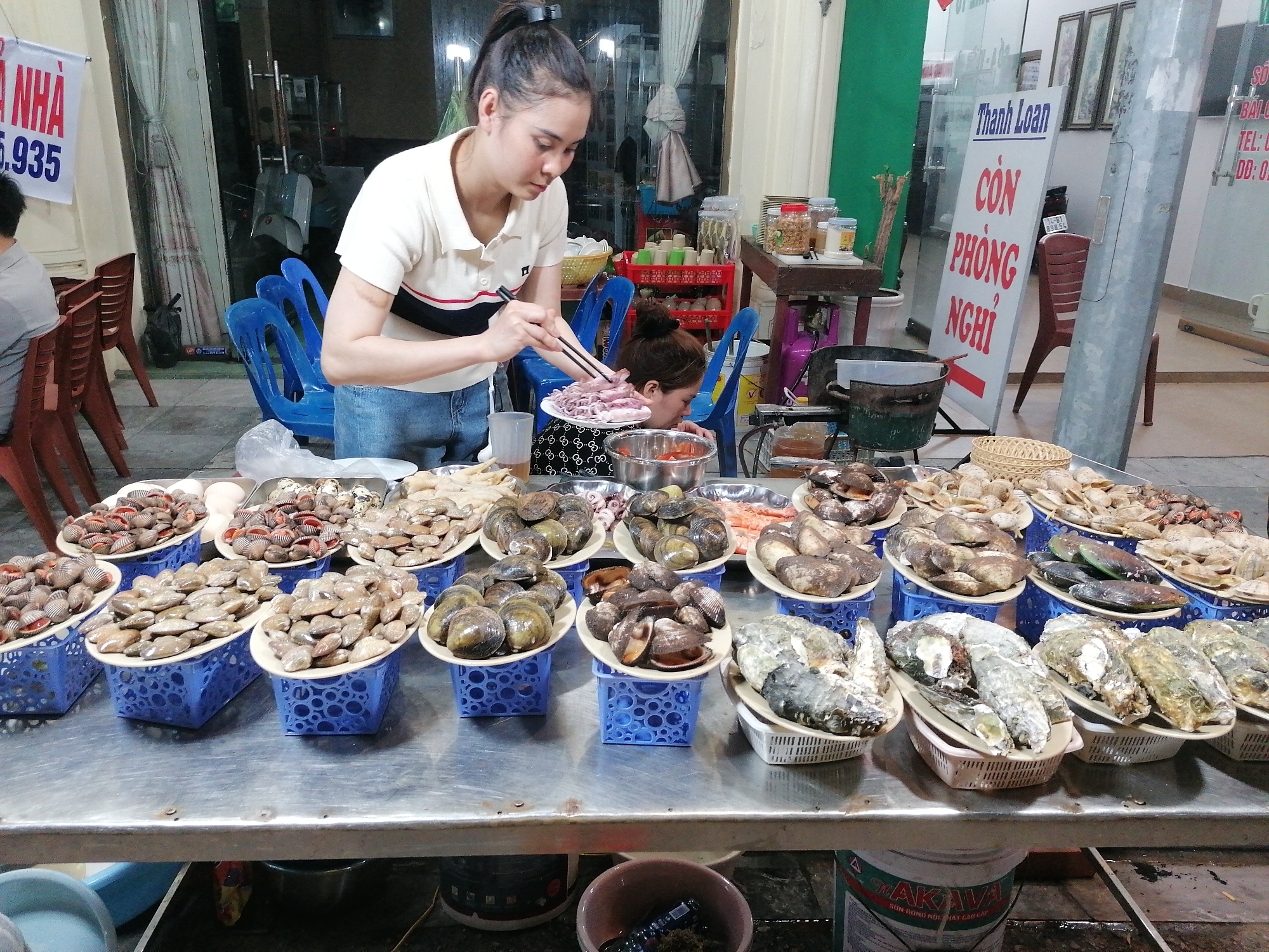 This media file captures an active seafood stall or restaurant at night, likely in Bai Chay Ward, Vietnam, given the provided context and visible text. The scene is illuminated by artificial lights, creating a warm, bustling atmosphere.
In the foreground, a young woman with her hair tied back, wearing a cream polo shirt with black and red stripes and jeans, is meticulously arranging raw squid with chopsticks onto a white plate. Behind her, another woman is partially visible, leaning over a table, possibly engaged in food preparation.
The central feature of the image is a long, metal display table laden with numerous plates and baskets filled with various types of fresh seafood. These include diverse shellfish like clams, oysters, mussels, scallops, and possibly some shrimp, all arranged neatly on white plates that are elevated by blue plastic baskets.
In the background, red and blue plastic chairs are visible, suggesting a seating area for customers. Further back, cooking equipment such as pots and gas canisters can be seen, indicating food is prepared on-site. A scooter is partially visible through a glass door.
Several signs with Vietnamese text are present. On the left, a red and white banner shows "NHÀ" and "935". A prominent blue and white sign in the middle reads "Thanh Loan CÒN PHÒNG NGHỈ", indicating "Thanh Loan" (likely a business name) and "Rooms Available", suggesting the food stall might be associated with a guesthouse or small hotel. Another sign on the right lists "TEL:" and "DD:" with partial numbers like "898" and "0". At the bottom, a repurposed bucket displays "AKAVA" and "SƠN BÓNG NỘI THẤT CAO CẤP" (High-grade interior gloss paint).
The overall impression is a vibrant, informal eatery specializing in fresh, likely cooked-to-order, seafood.