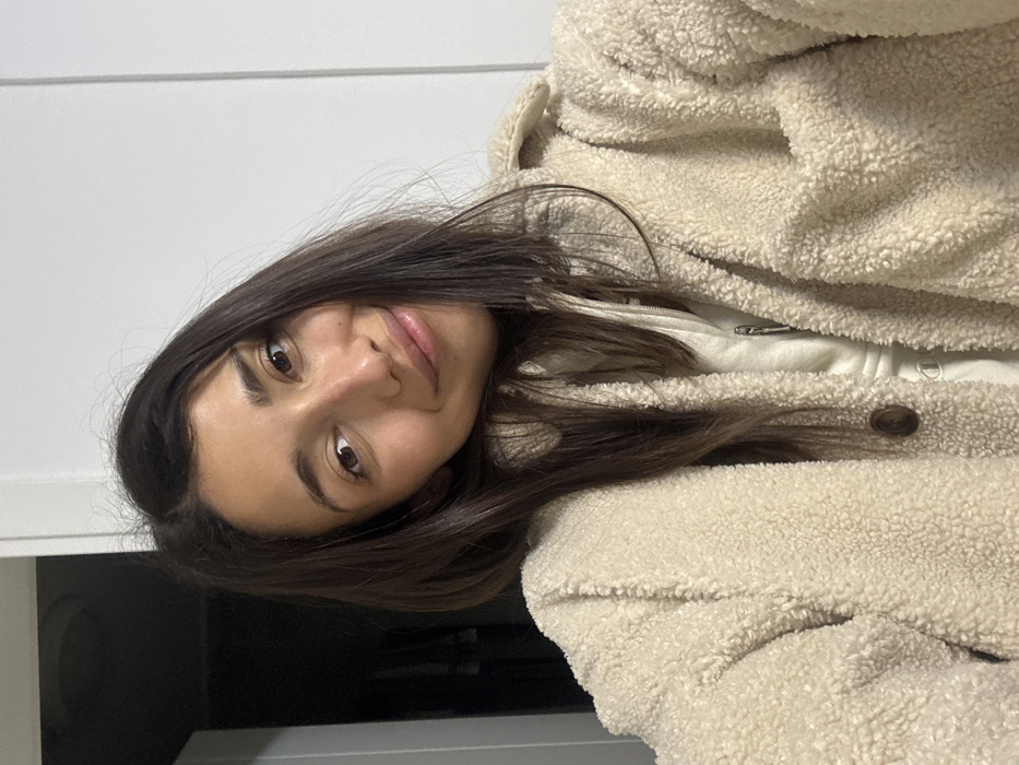 A young woman with long brown hair poses for a selfie. She wears a cream-colored, plush jacket over a lighter-colored top. Her expression is neutral. The background is a neutral-colored wall or surface.