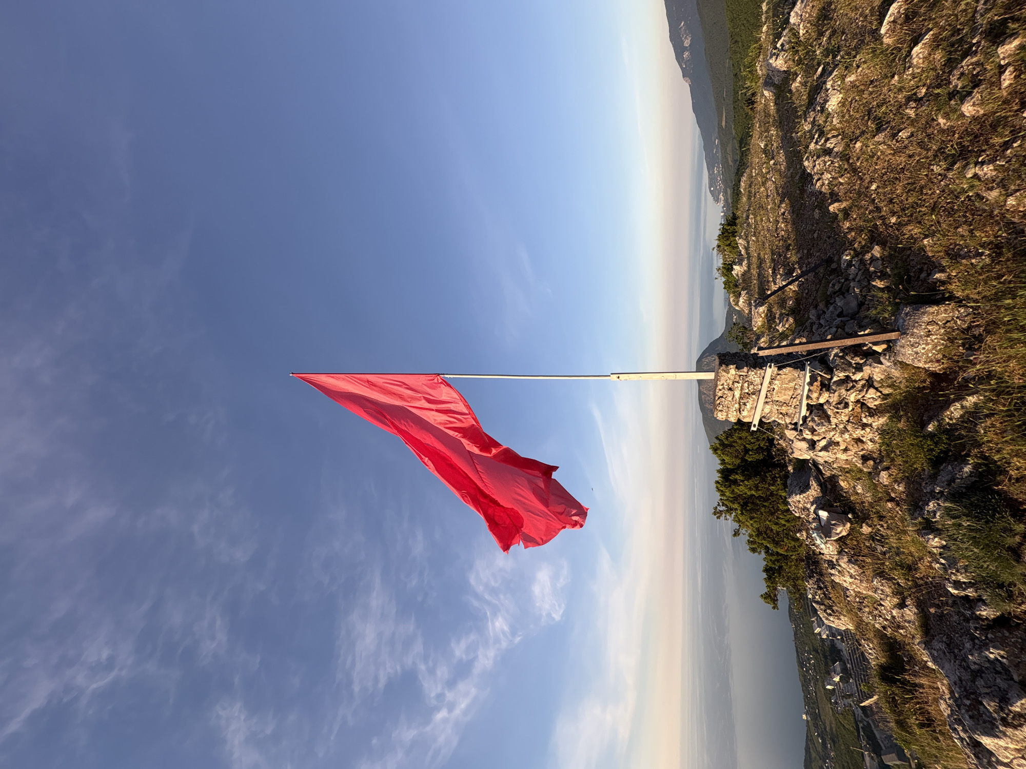 A bright red rectangular flag, unfurled and exhibiting undulations from wind, stands on a metallic flagpole atop a rocky summit. The flagpole is anchored in a base constructed from stacked stones and integrated metal supports. The immediate foreground features rugged terrain with light-colored rocks, exposed soil, and sparse patches of dry, yellowish-green grass. In the middle ground, to the left, a coastal area is visible with numerous white structures, indicative of buildings, along a descending slope. A vast, calm body of water extends across the horizon, meeting a hazy distant landmass. To the right, forested mountain ranges recede into the background. The sky is clear blue, with scattered, wispy cirrus clouds present predominantly in the upper-left quadrant. The lighting conditions suggest either early morning or late afternoon. No individuals are present in the scene. The location is a coastal mountainous region in Ukraine, near an unspecified city.