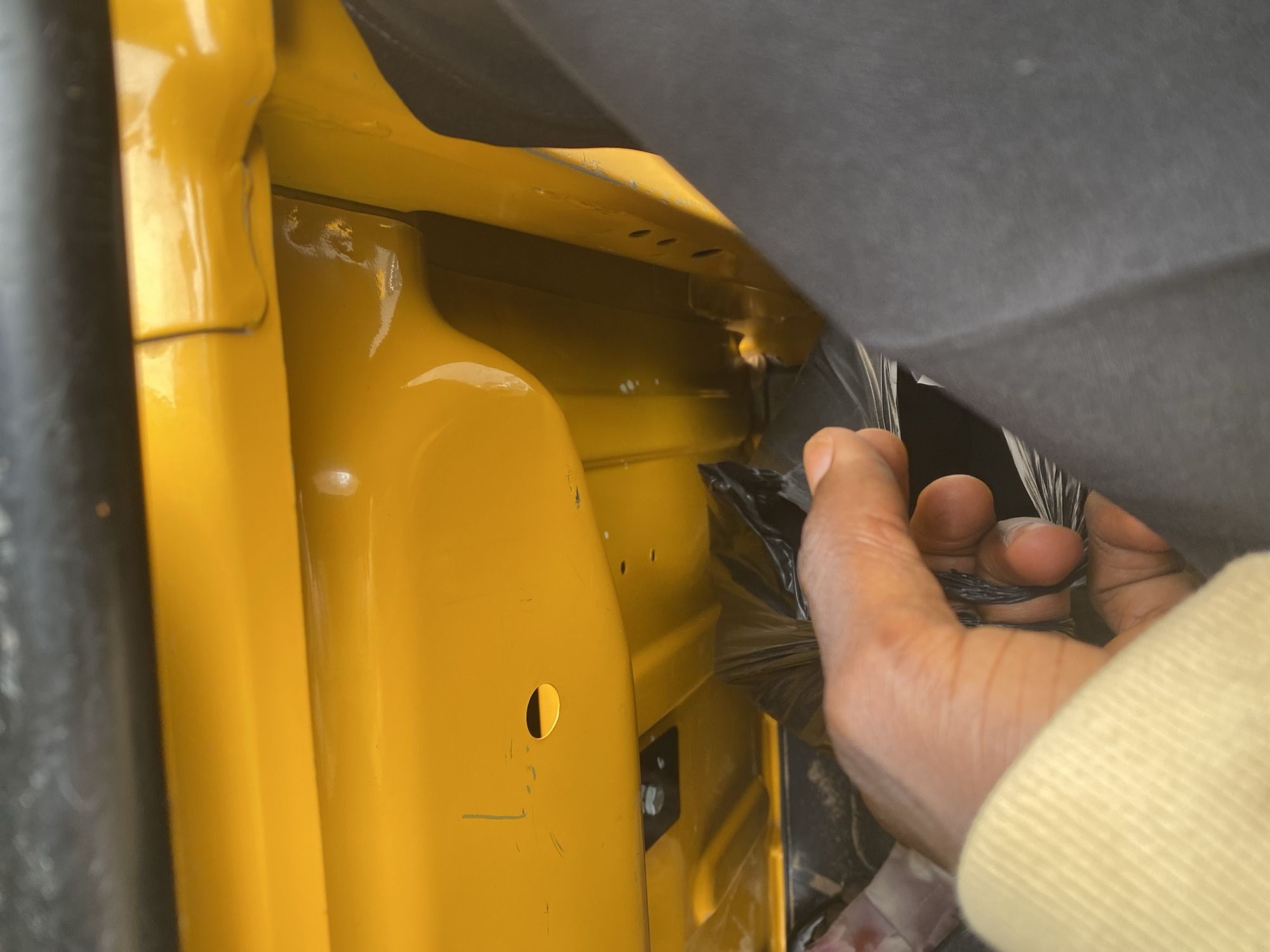 The image shows a close-up view of a yellow, potentially metallic, surface with some scuff marks and a small circular hole. A person's hand, with dark skin and a yellow sleeve, is reaching into a black plastic bag. The context suggests this might be inside a vehicle or some sort of enclosure with yellow painted metal. The location is specified as Jalingo, Nigeria.