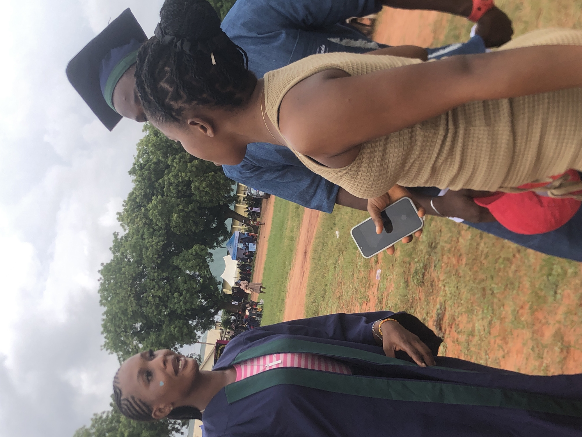 The image shows a group of people outdoors in what appears to be a celebratory setting. A woman wearing a graduation gown with green trim and a pink and white striped shirt underneath is smiling and looking towards the left side of the frame. To the right, a woman wearing a textured beige top is holding a smartphone. Behind them is a man wearing a graduation cap. The environment includes trees, grass, and what seems to be a gathering of people further in the background. The setting is in the city of Nsugbe, Nigeria.